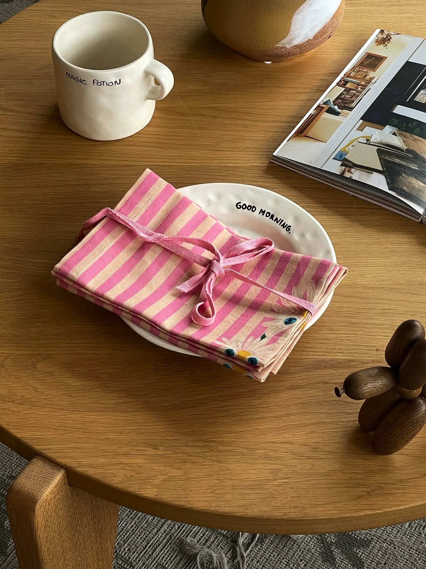An Anna + Nina Trippy Flower Napkin, Set of 2, featuring pink stripes and a floral motif, is tied with a ribbon on a white GOOD MORNING plate beside a MAGIC POTION cup and an open magazine for that perfect spring vibe.