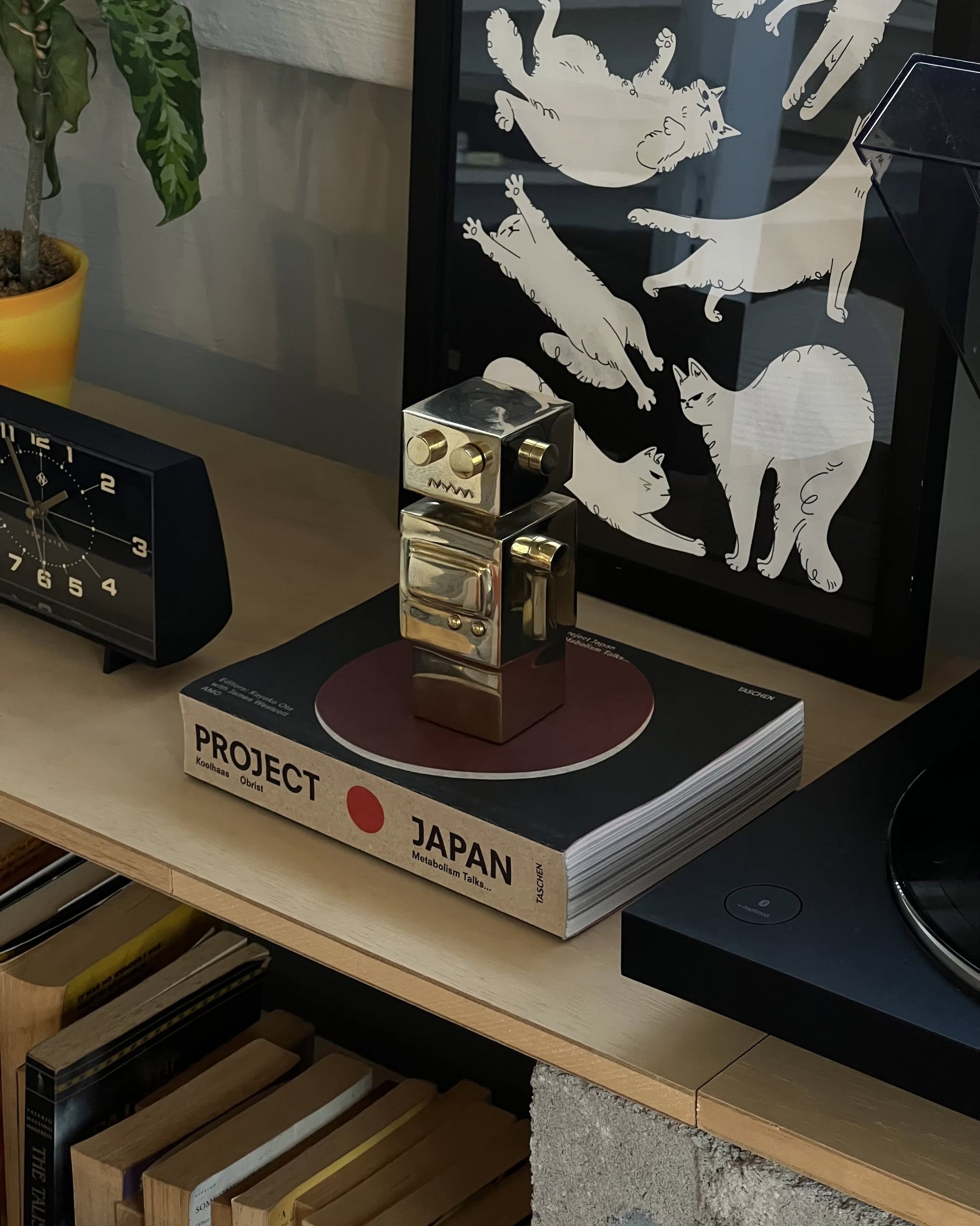 The Anna + Nina Back to the Future Figure stands on a book titled Project Japan beside a black clock and framed playful white cat artwork, all arranged on a shelf with books and a potted plant.