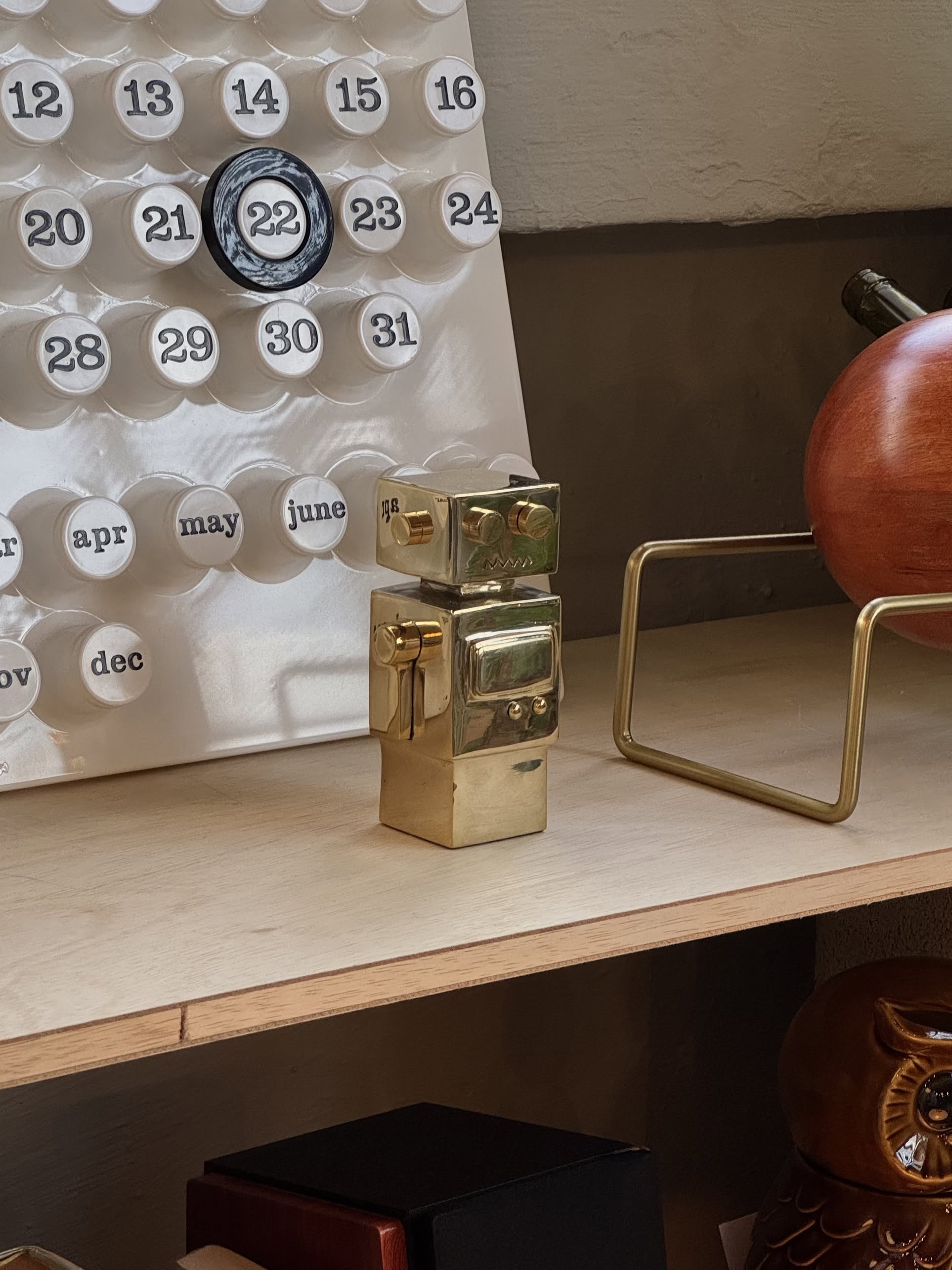 The Anna + Nina Back to the Future Figure, a small brass piece, is placed on a wooden shelf next to a white perpetual calendar and other decor. Behind it are a brown object and part of a gold-finished metal frame.