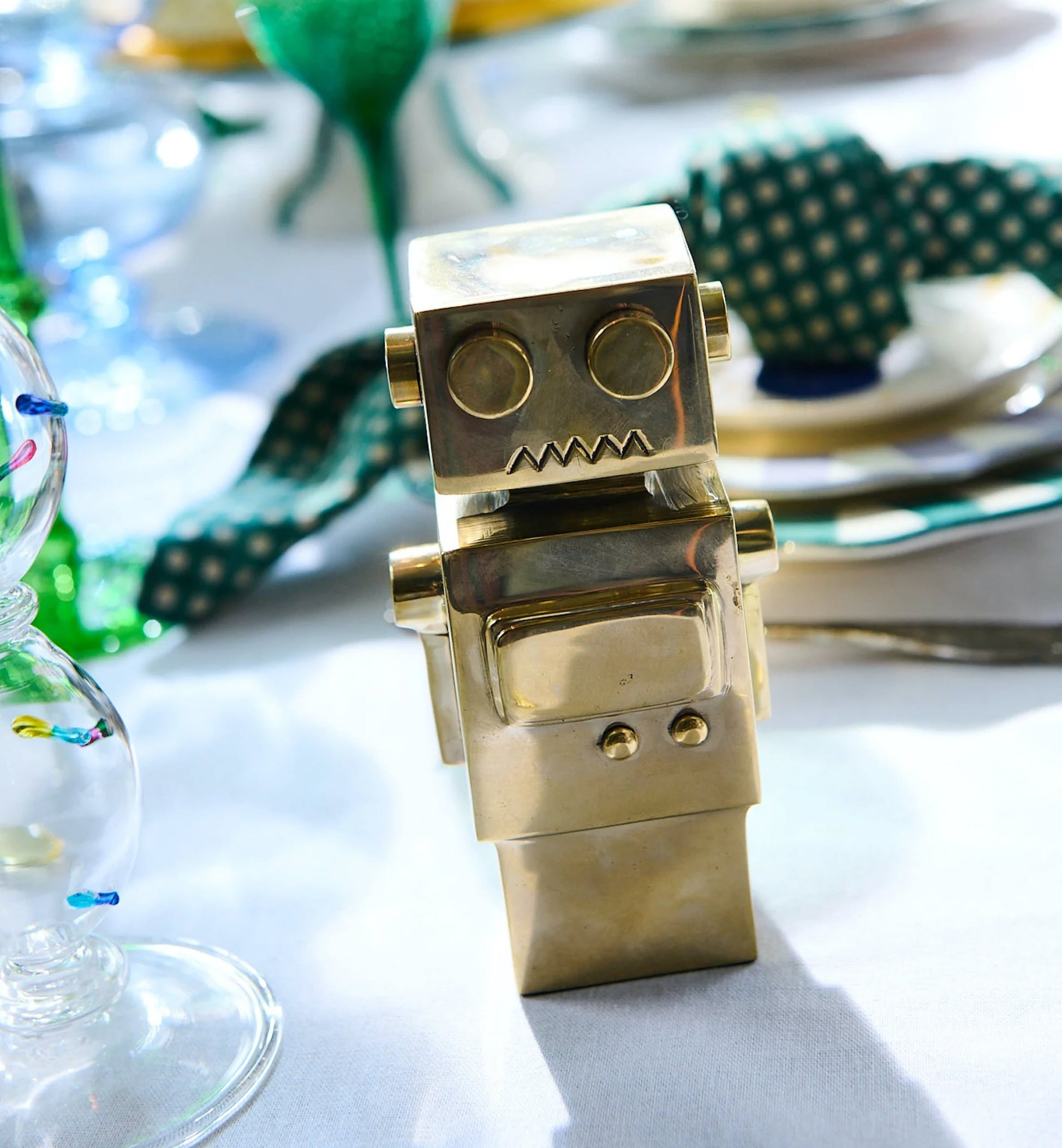 The Anna + Nina Back to the Future Figure, a small robot-inspired gold figurine with a square head and body, stands on a table set with plates, glasses, and a green polka-dot napkin in the background.