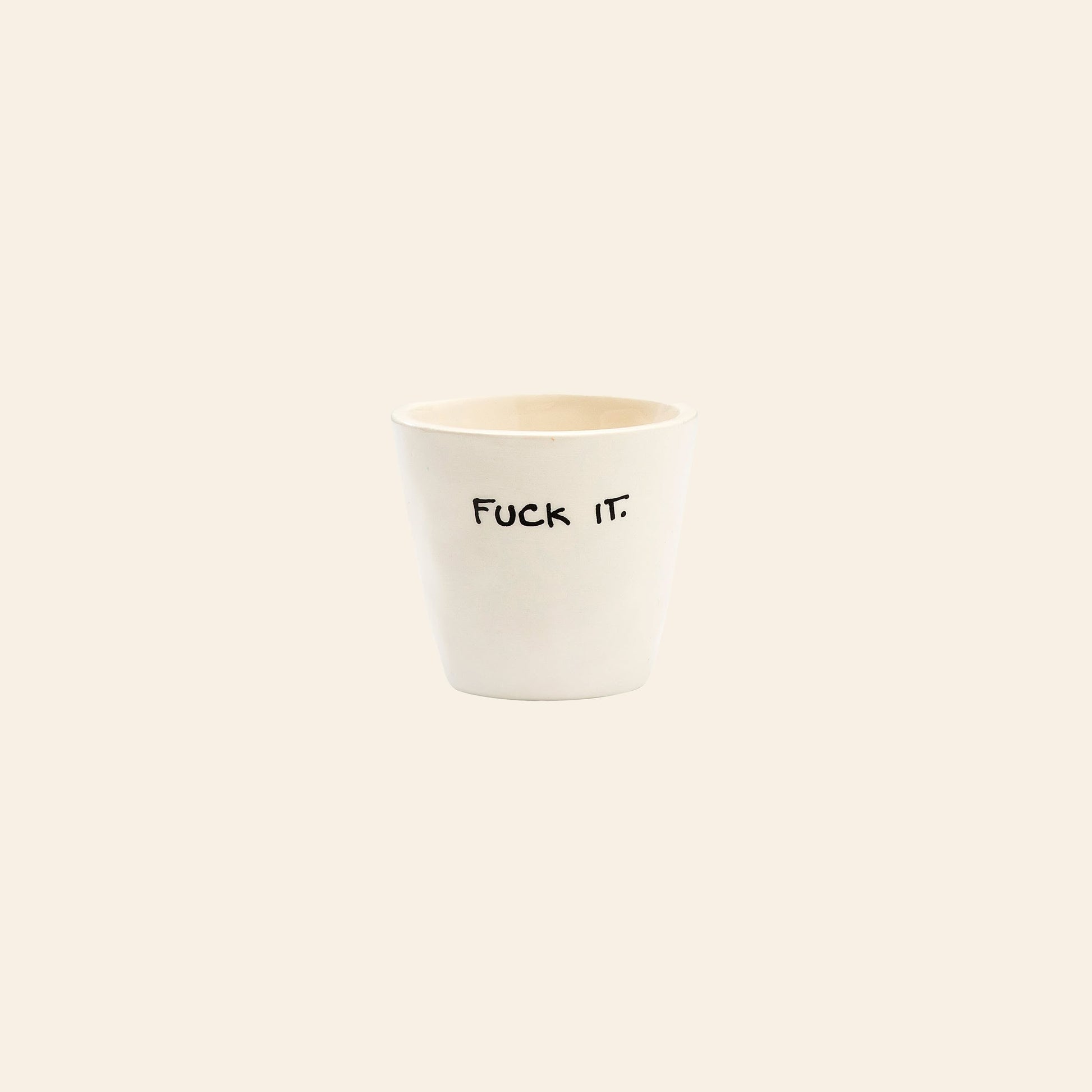The Anna + Nina Fuck It Espresso Cup by Anna + Nina is a white ceramic cup with Fuck It. in black text, set against a beige background—perfect for adding a playful touch of coffee humor to your daily routine.