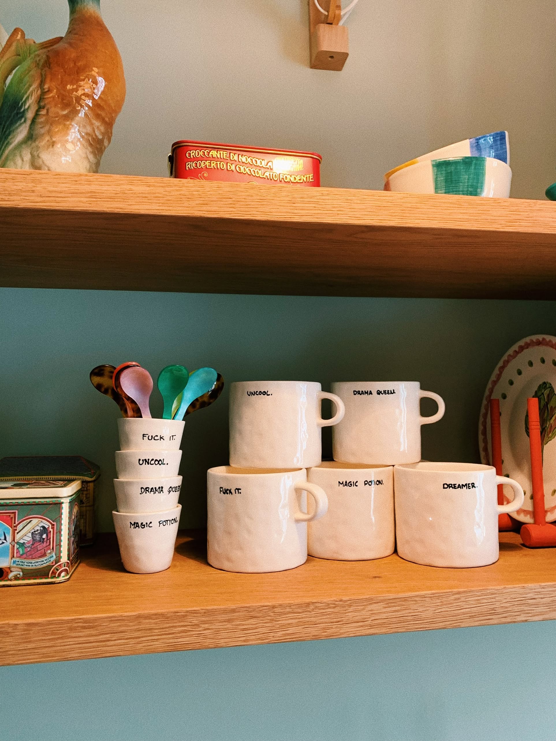 Two wooden shelves display assorted ceramic cups and mugs, like the Anna + Nina Fuck It Espresso Cup. Each mug or cup has handwritten labels such as Magic Potion, Dreamer, and Uncool. Decorative tins and plates add extra charm nearby.