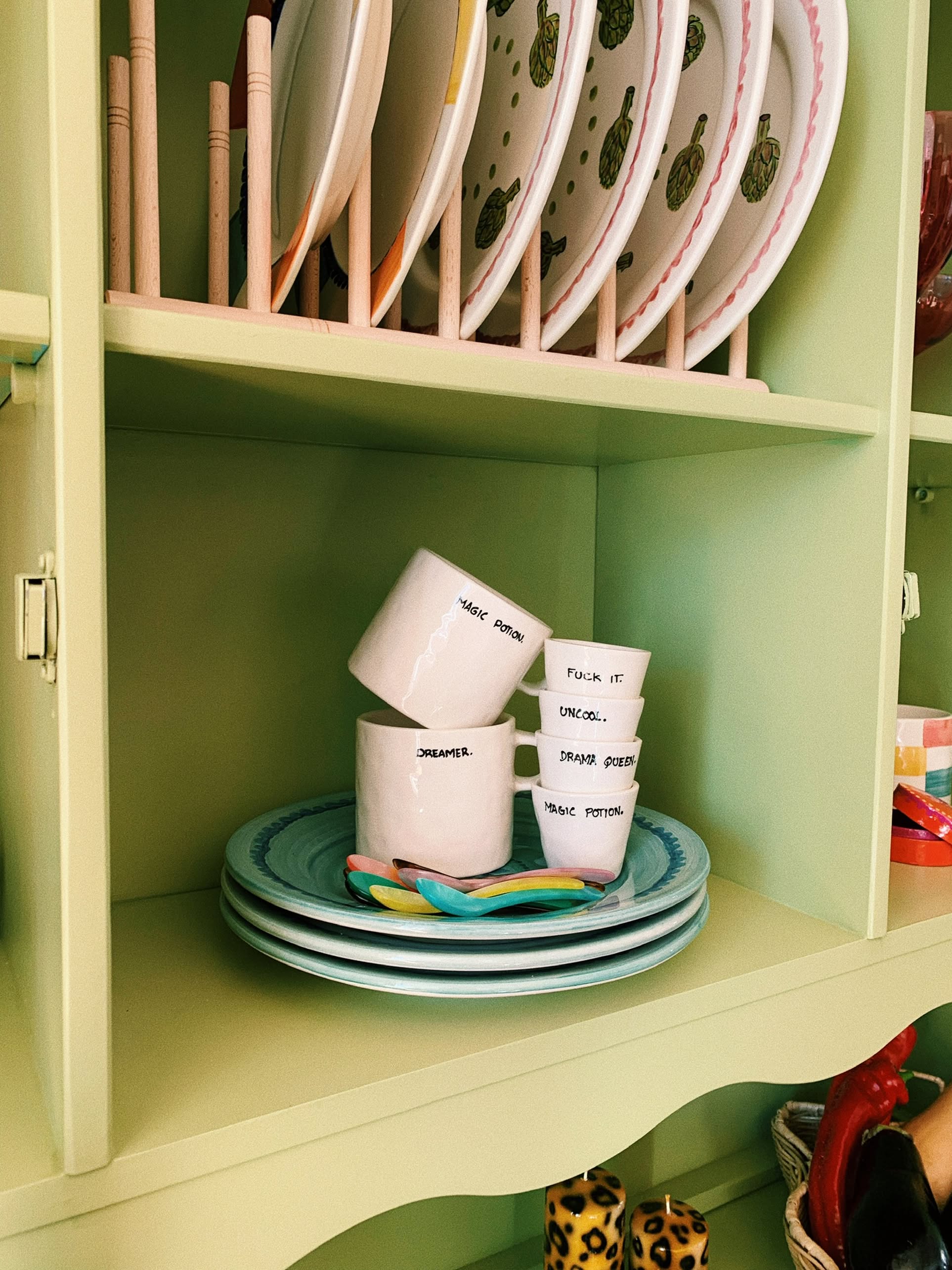 A pastel green shelf displays Anna + Nina Fuck It Espresso Cups, stacked teal plates, colorful turtle-design plates, a ceramic cup, three white mugs with coffee humor phrases, and two colorful spoons for a playful touch.