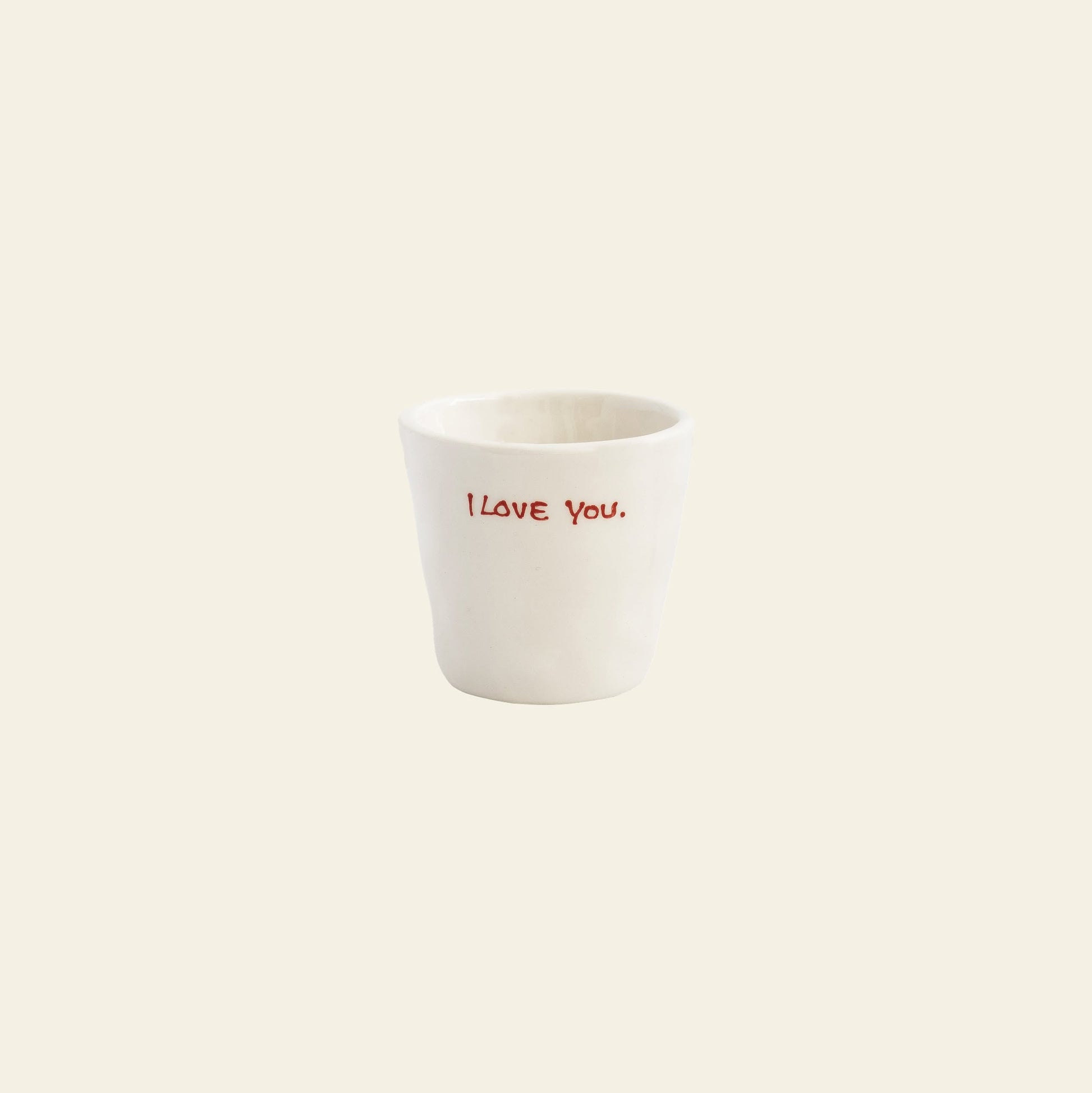 The Anna + Nina I Love You Espresso Cup is a white ceramic espresso cup with I love you. in red on the front, set against a light beige background—an ideal thoughtful gift from Anna + Nina.