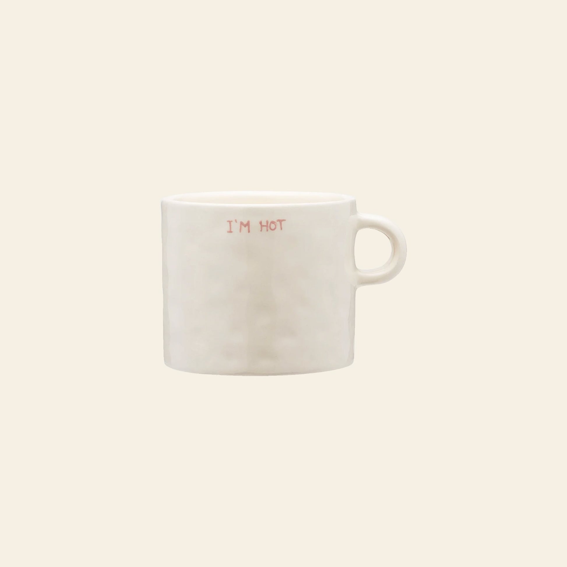 The Anna + Nina Im Hot Mug is a plain white ceramic mug with a handle, featuring IM HOT in small red letters on the front—ideal for hot drinks or gifting. Shown against a light beige background.