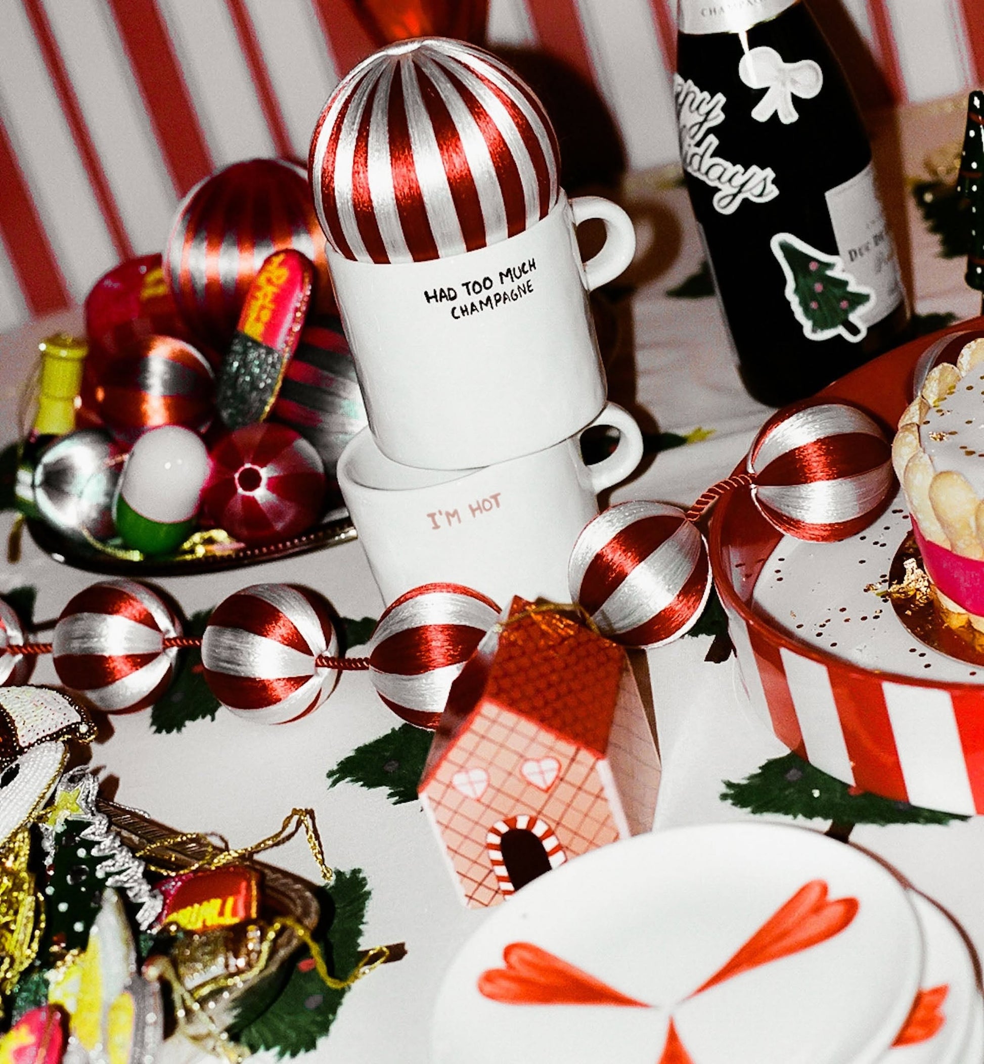 A festive table features red and white decorations, baubles, a gifting-ready Anna + Nina I’m Hot Mug filled with a hot drink, the HAD TOO MUCH CHAMPAGNE mug, a gingerbread house, bottle, and candy cane heart shapes on a plate.