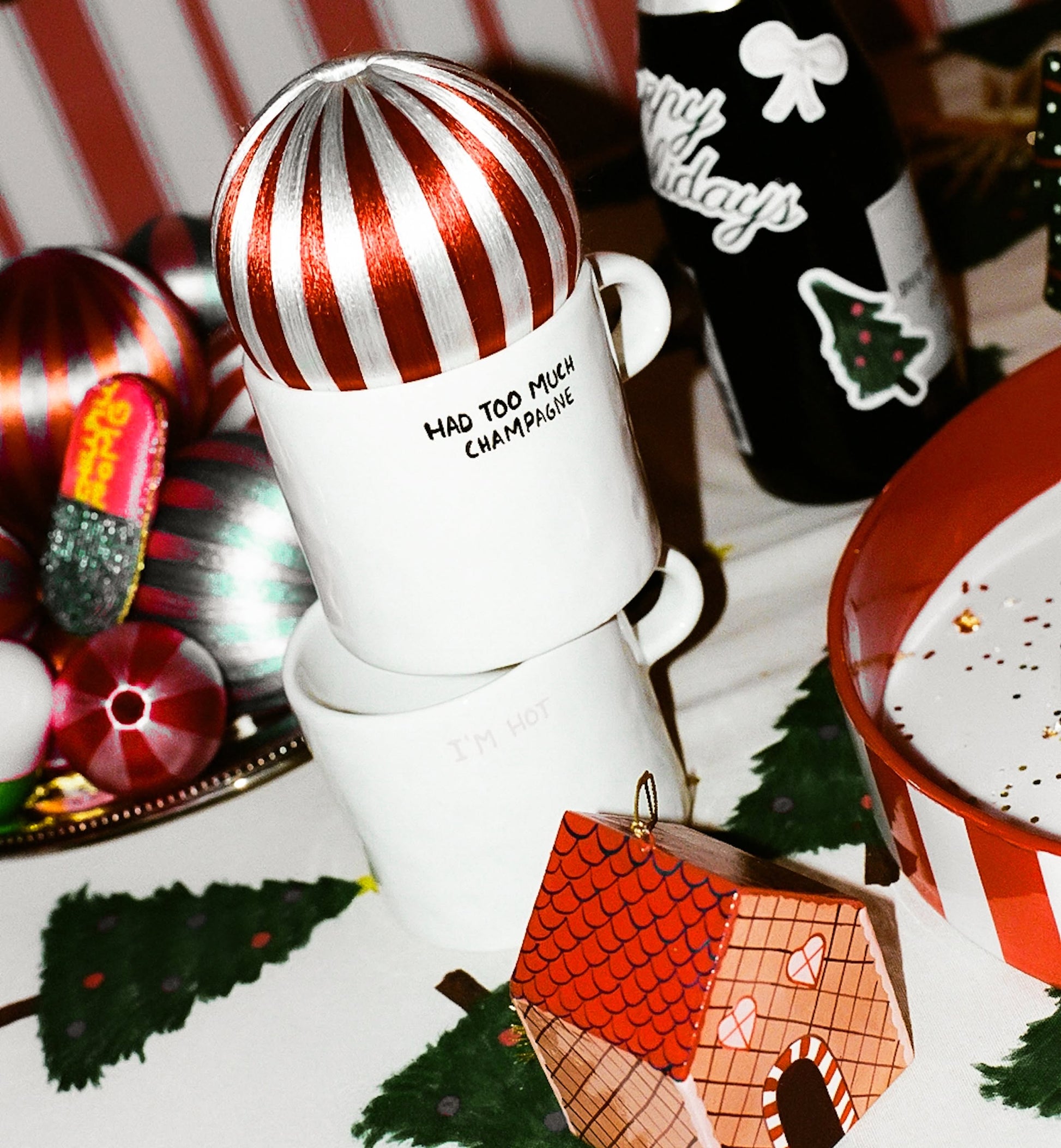 Two stacked Anna + Nina Im Hot Mugs sit beside hot drinks and a black Happy Holidays bottle, all surrounded by holiday décor—making this festive Anna + Nina setup perfect for gifting or cozy celebrations.
