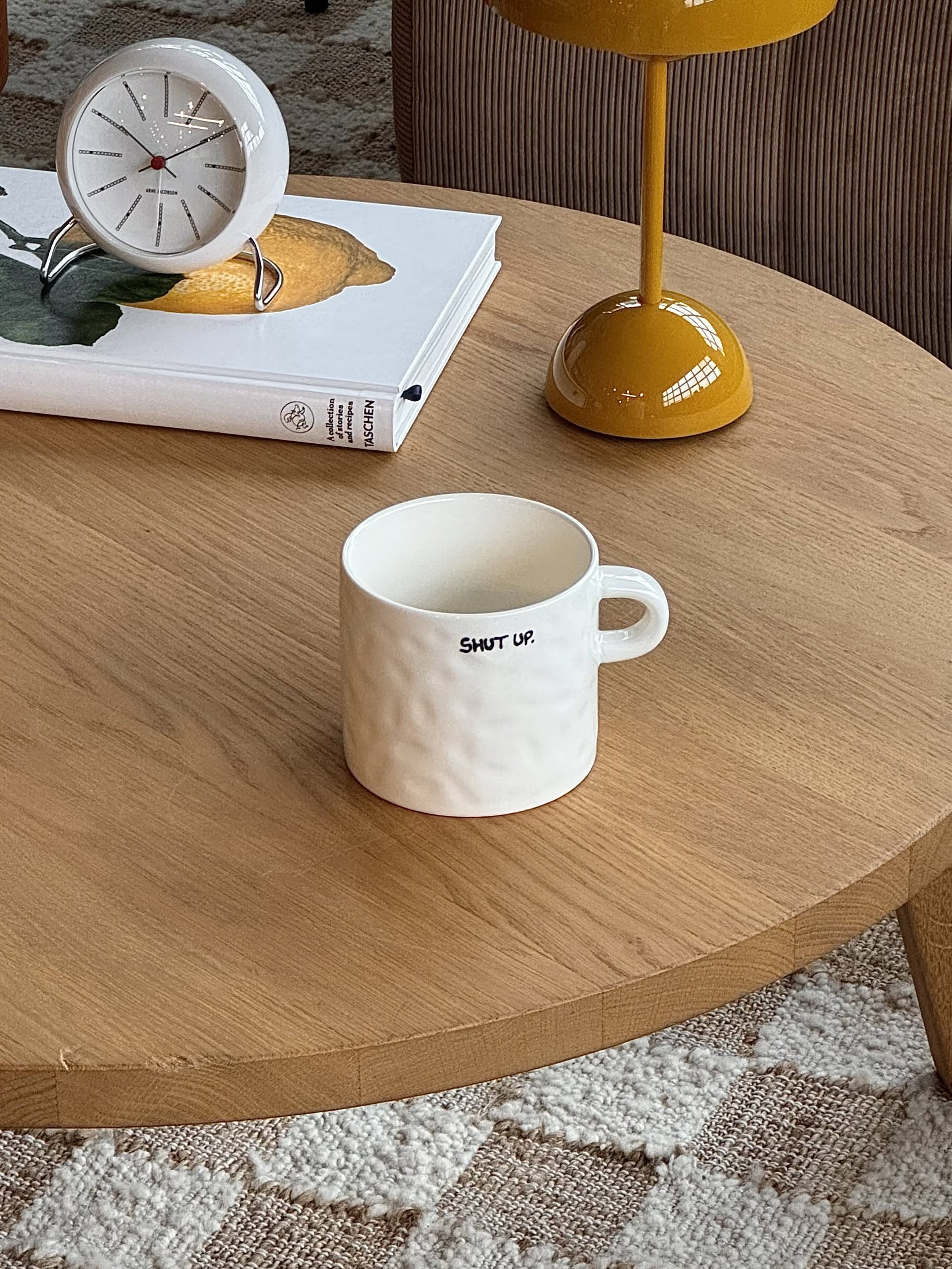 The Anna + Nina Shut Up Mug, made of white ceramic, rests on a wooden coffee table next to a yellow lamp, a lemon-covered book, and a white clock, with part of a patterned rug visible beneath the table.