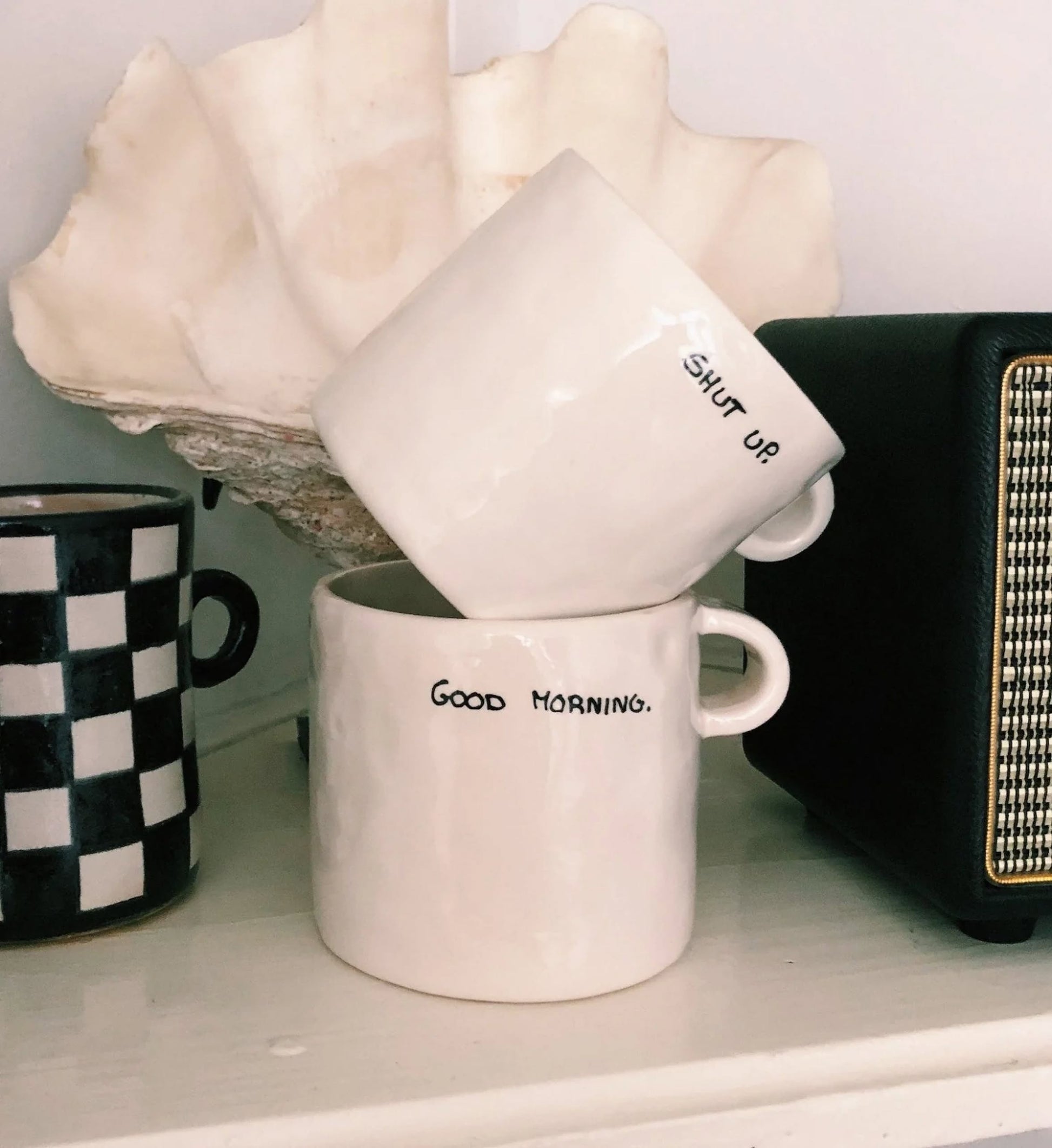 Two white ceramic mugs—a Shut Up Mug by Anna + Nina and another reading GOOD MORNING—are stacked on a shelf. Nearby are a black-and-white checkered mug, a large shell, and part of a speaker, creating a quirky scene for caffeine lovers.