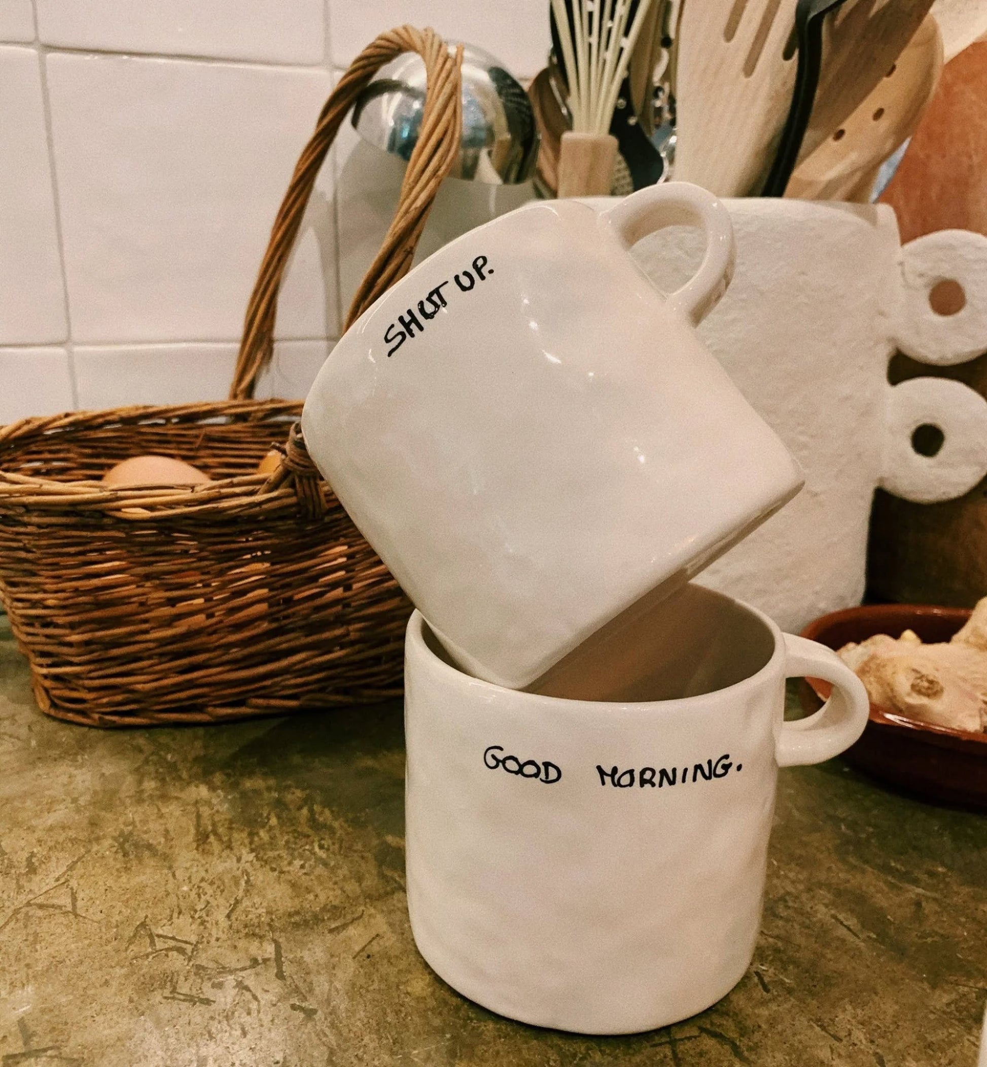 Two white Anna + Nina ceramic mugs are stacked on a kitchen counter. The top, the Anna + Nina Shut Up Mug, reads SHUT UP, while the bottom one says GOOD MORNING. Kitchen utensils and a wicker basket are in the background.