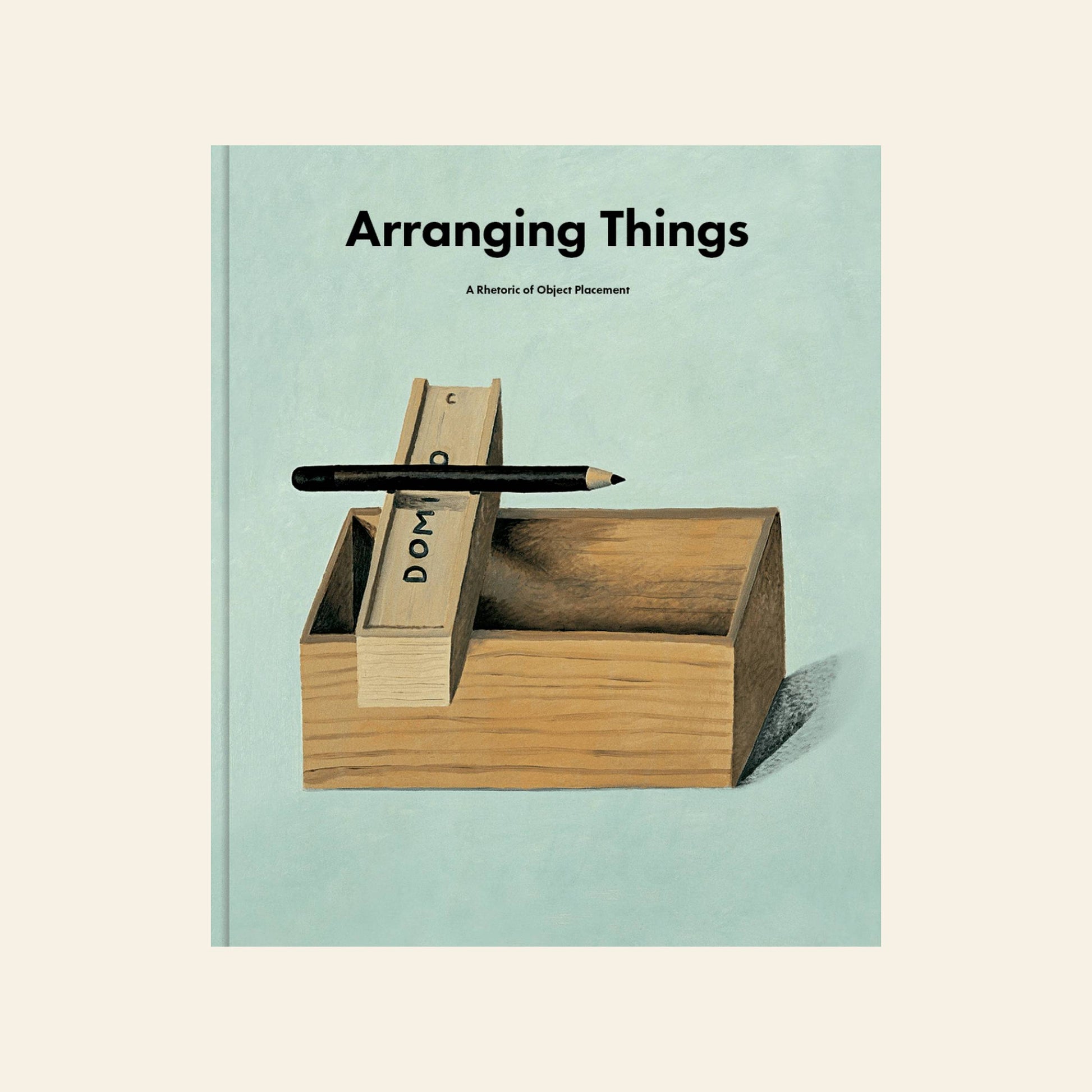 The cover of Apartamento Arranging Things: A Rhetoric of Object Placement by Apartamento shows a wooden box with dominoes and a pencil on top, reflecting its design ethos against a soft, light background.
