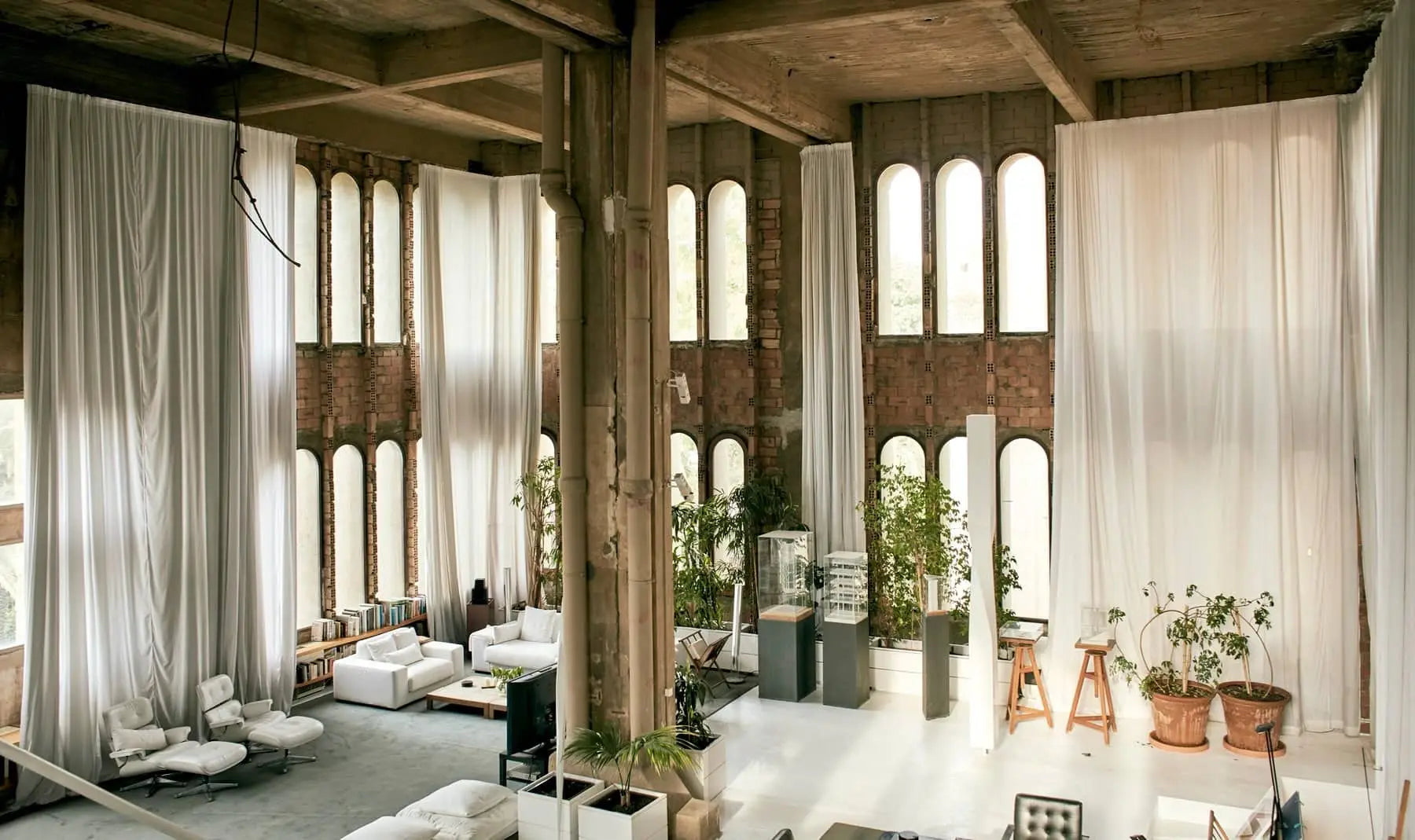 The Apartamento La Fábrica by Apartamento features an industrial style inspired by Ricardo Bofill, with tall arched windows, exposed brick, large columns, white curtains, modern furnishings, bookshelves, and potted plants for a bright look.