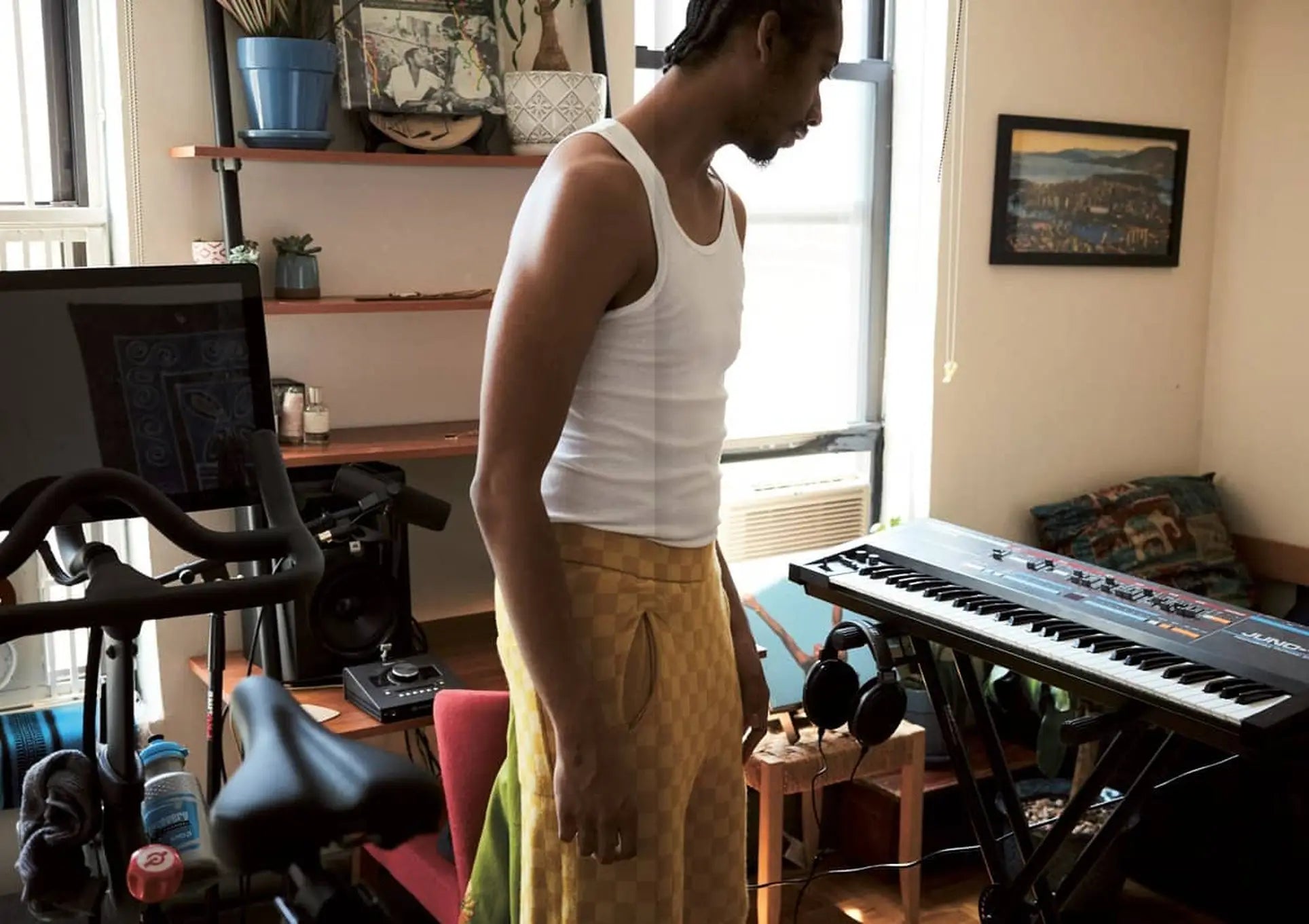 A person in a white tank top and yellow patterned pants stands in a bright room with musical gear, art inspired by Niki de Saint Phalle, an exercise bike, and Apartamento Magazine Issue #28 by Apartamento.