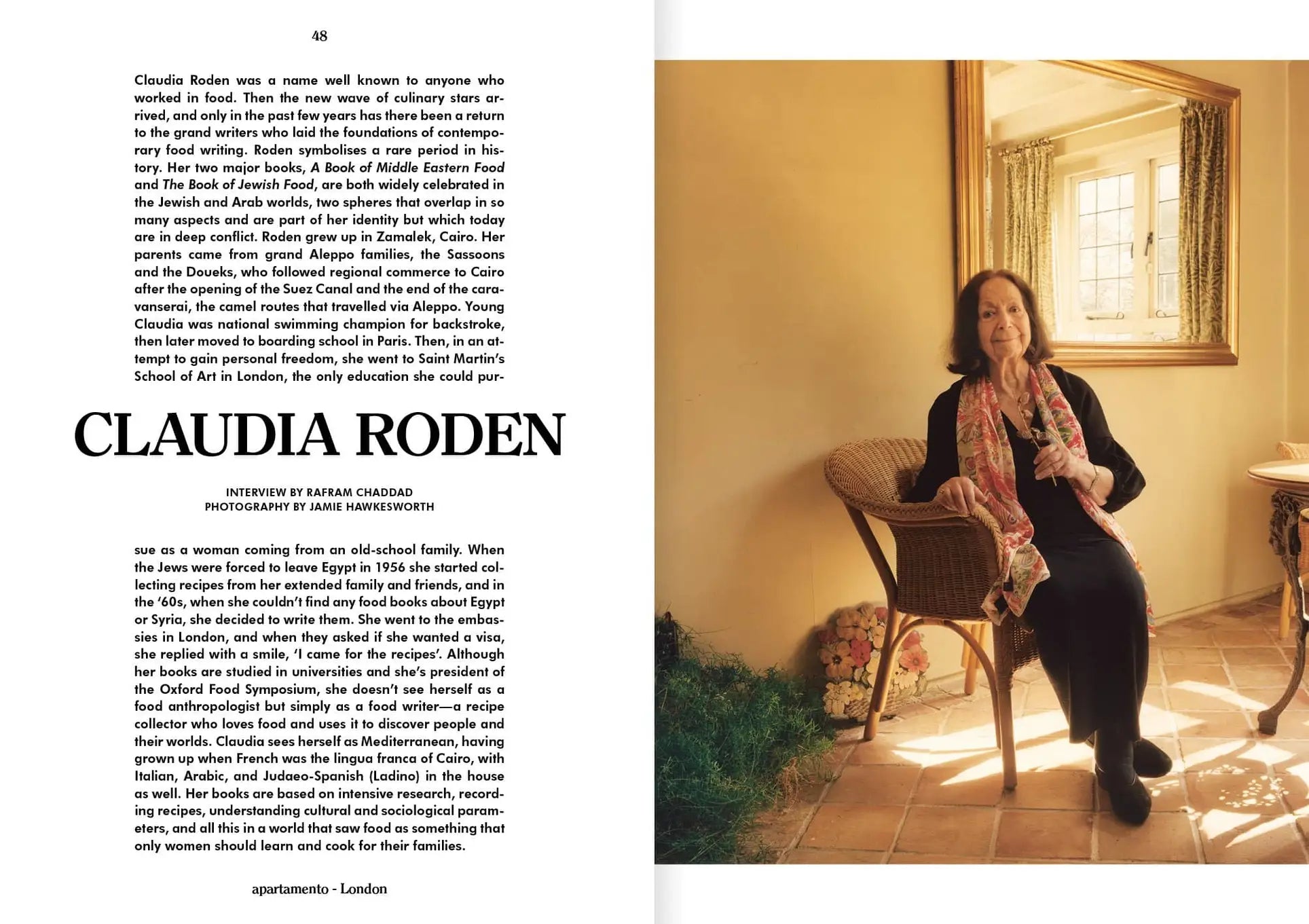 A woman with long hair sits on a wooden chair in a sunlit room at Siwa Oasis, holding a flower—a serene scene from Spring/Summer 2022, captured in Apartamento Magazine Issue #29 by Apartamento. Lush plants flourish outside the window.
