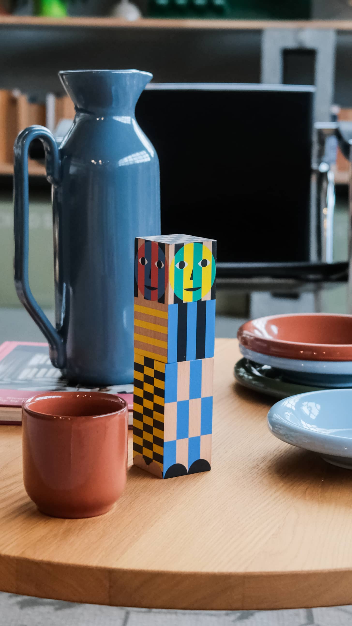 The Areaware Everybody Grinder by Areaware stands on a round wooden table, surrounded by ceramic plates, a mug, and a tall blue pitcher—ready to add spice and flavor to the kitchen scene with its colorful geometric design.