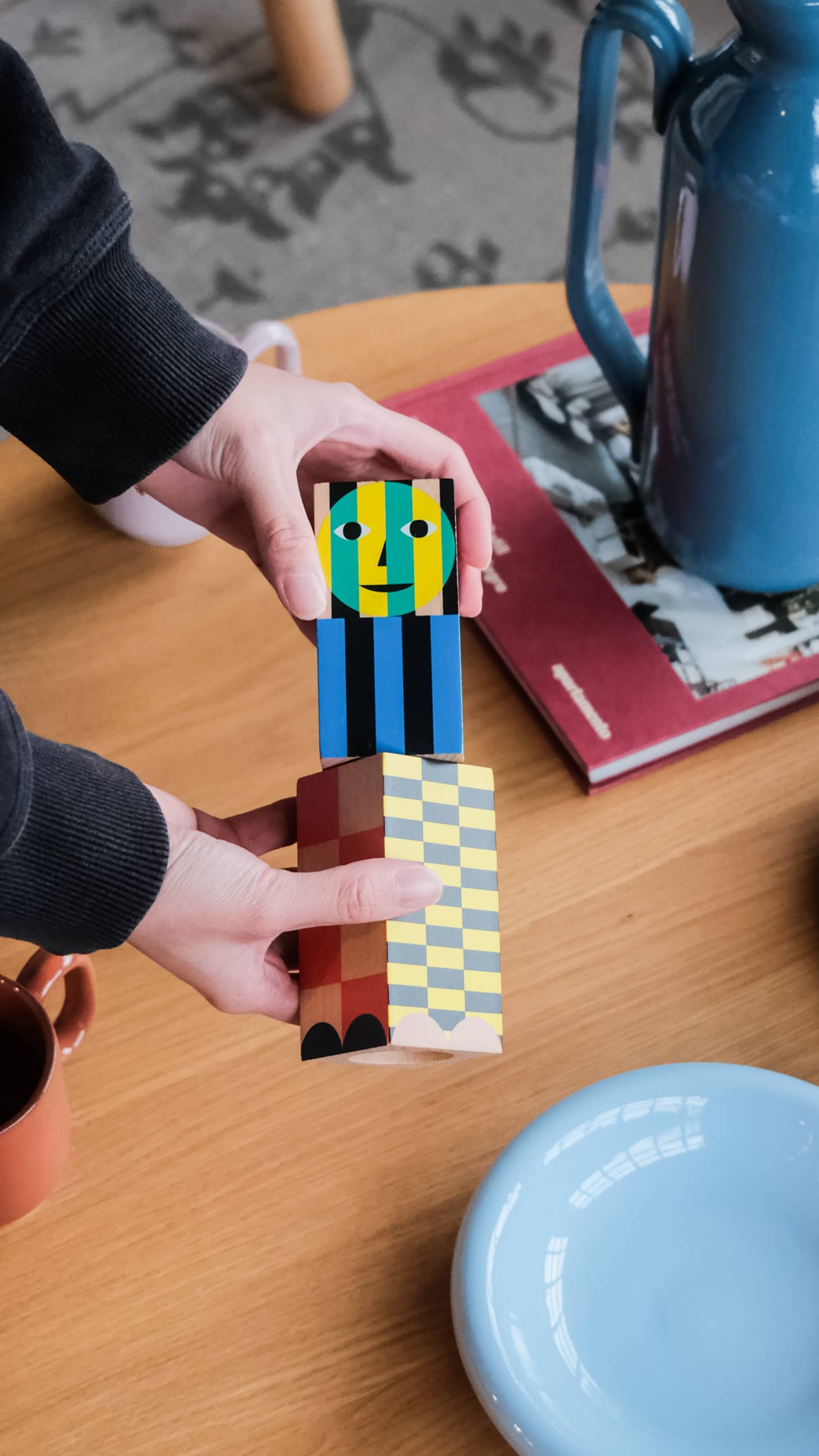 A person holds two colorful Areaware Everybody Grinders above a wooden table set with a blue jug, blue plate, red book, brown cup, and kitchen tool for adding spice.