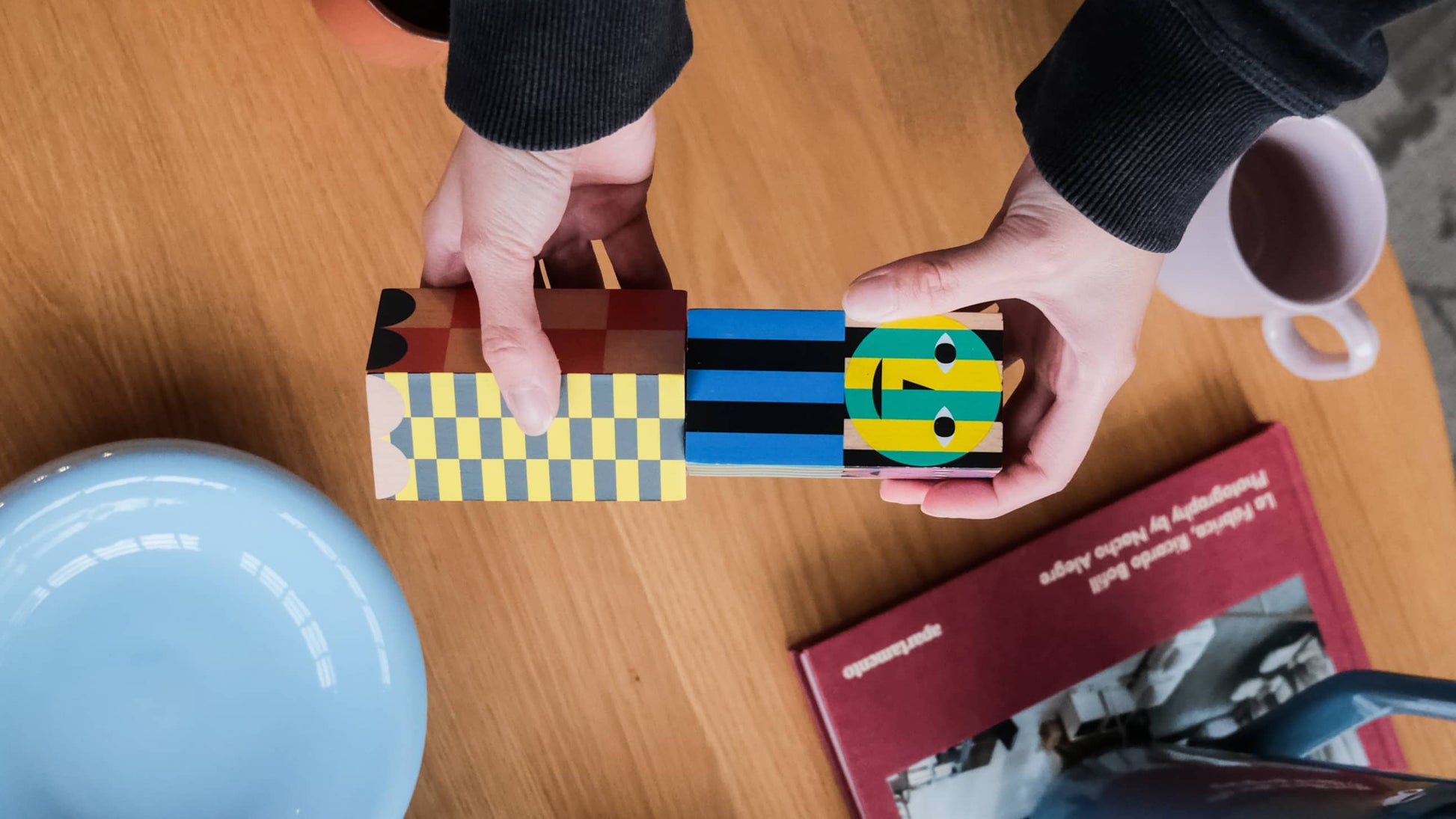 A persons hands hold colorful wooden blocks shaped like people over a wooden table with a blue plate, pink cup, red book, and the Areaware Everybody Grinder by Areaware adding spice and style to the scene.