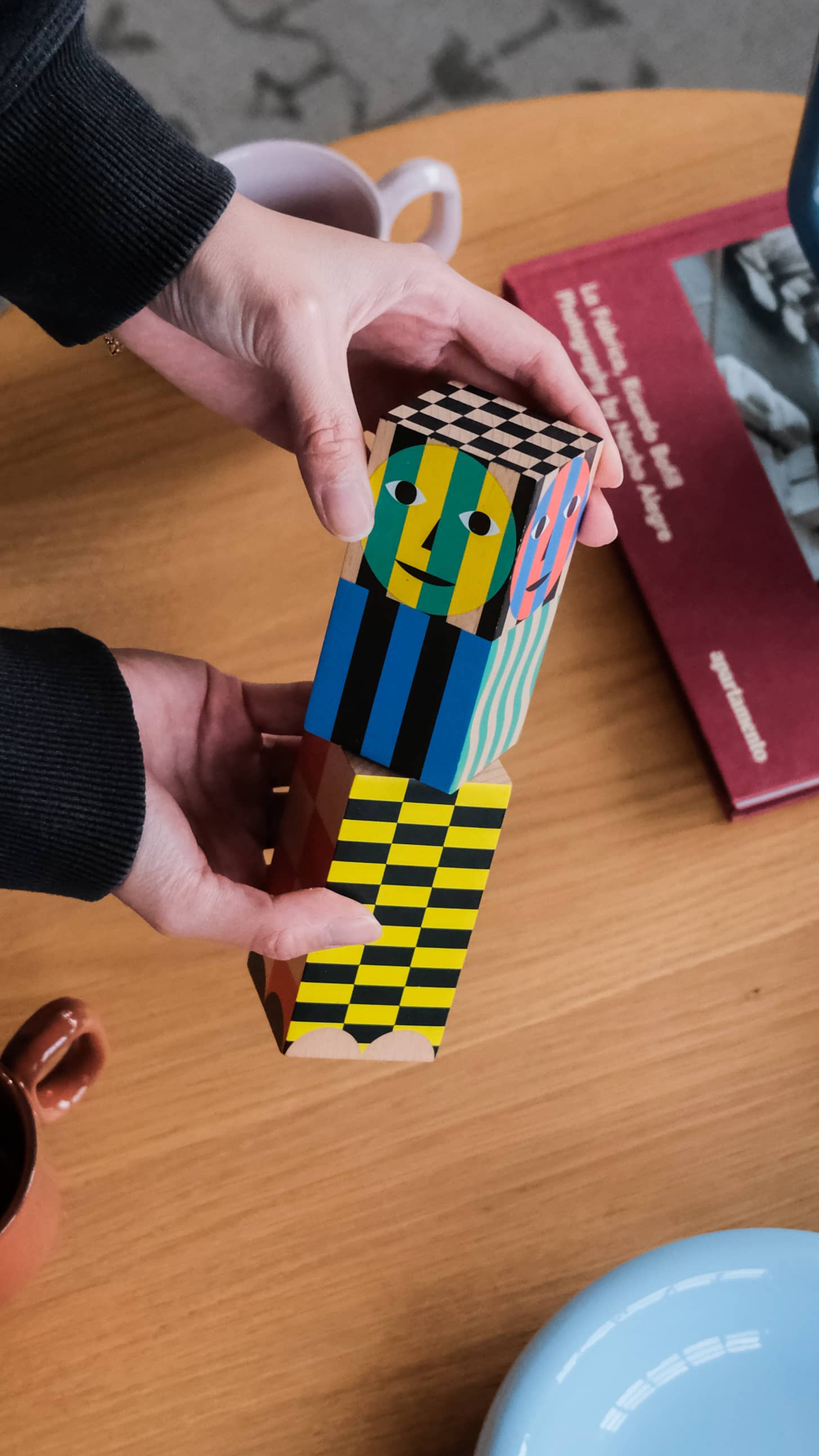 Two hands hold the Areaware Everybody Grinder by Areaware, a colorful, patterned grinder stacked to resemble a robot. Stripes, checks, and a face decorate it. A red book and coffee mug rest on the wooden table below.