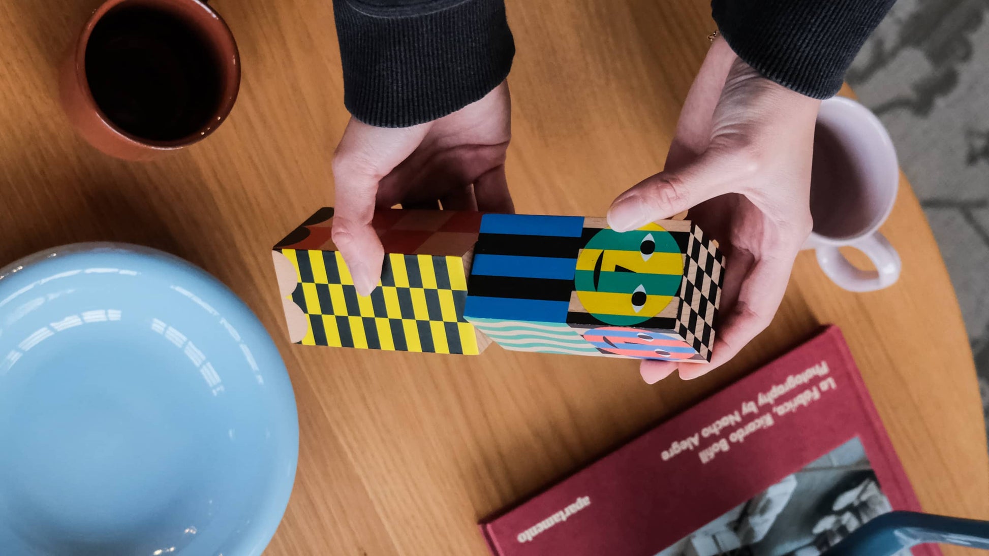 A person arranges colorful, patterned blocks with face designs on a wooden table, surrounded by a blue bowl, a red book, two mugs, and the Areaware Everybody Grinder by Areaware that adds spice and flavor to the vibrant scene.