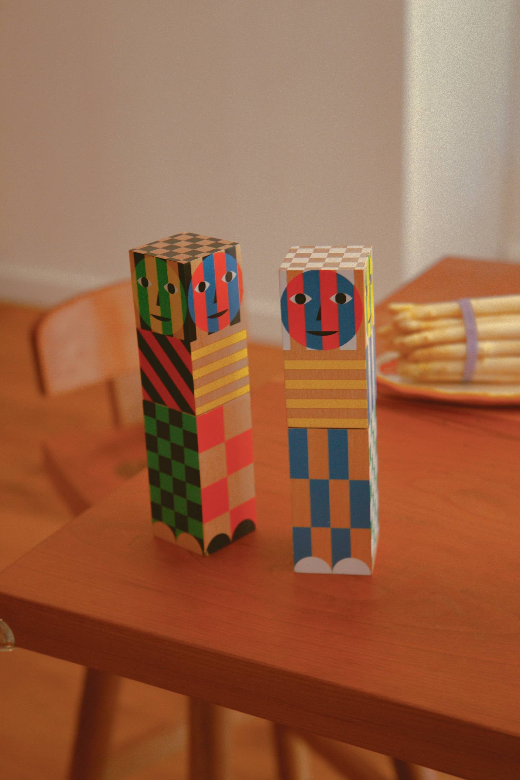 Two colorful Areaware Everybody Grinders with geometric faces stand on a wooden table, adding flair to the scene; a plate of white asparagus and a wooden chair are visible in the background.