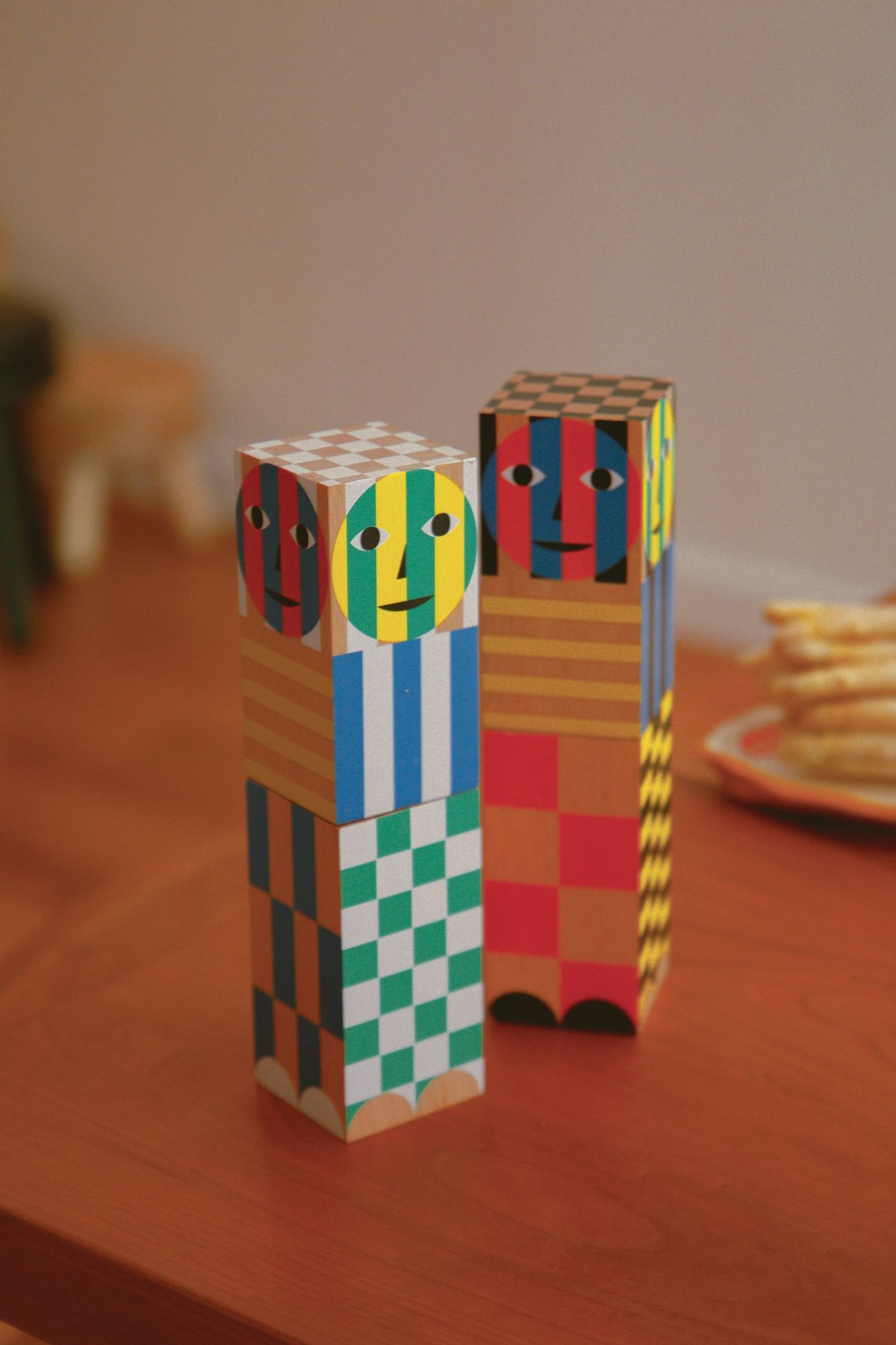 Two Areaware Everybody Grinders by Areaware, featuring vibrant geometric patterns and faces, stand upright on a wooden table. A blurred plate of snacks and kitchen tools in the background add to the cozy household atmosphere.