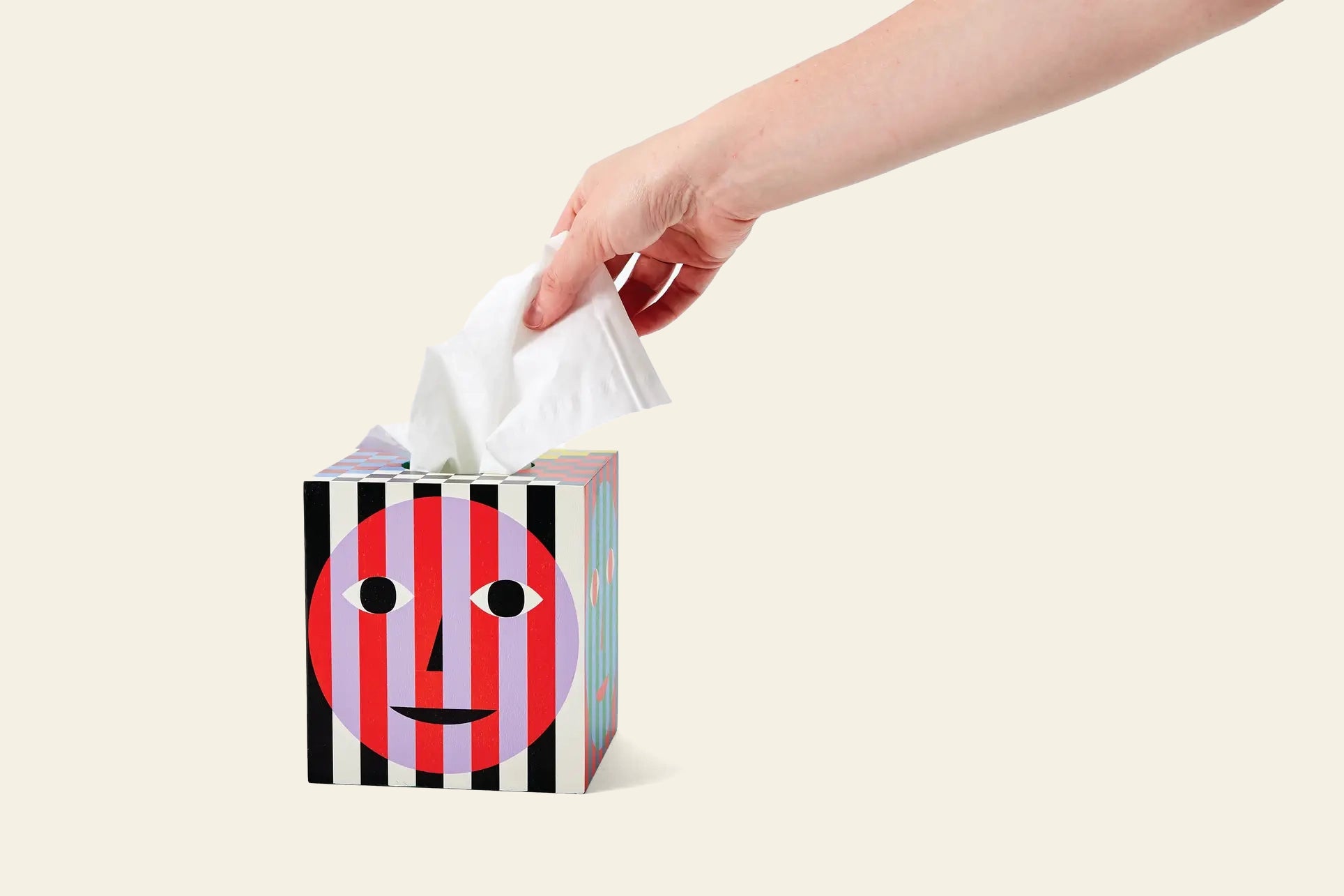 A hand pulls a tissue from the Areaware Everybody Tissue Box by Areaware, featuring a smiling face and bold vertical black, white, and red stripes on a plain light background.