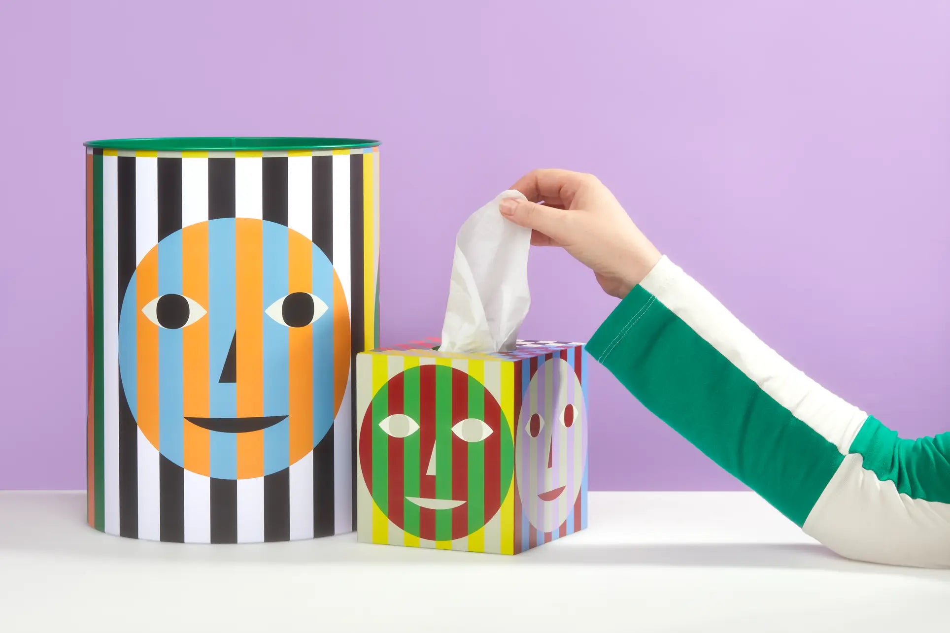 A hand pulls a tissue from an Areaware Everybody Tissue Box, its colorful striped design matching a larger cylindrical container next to it on a white surface against a purple background.