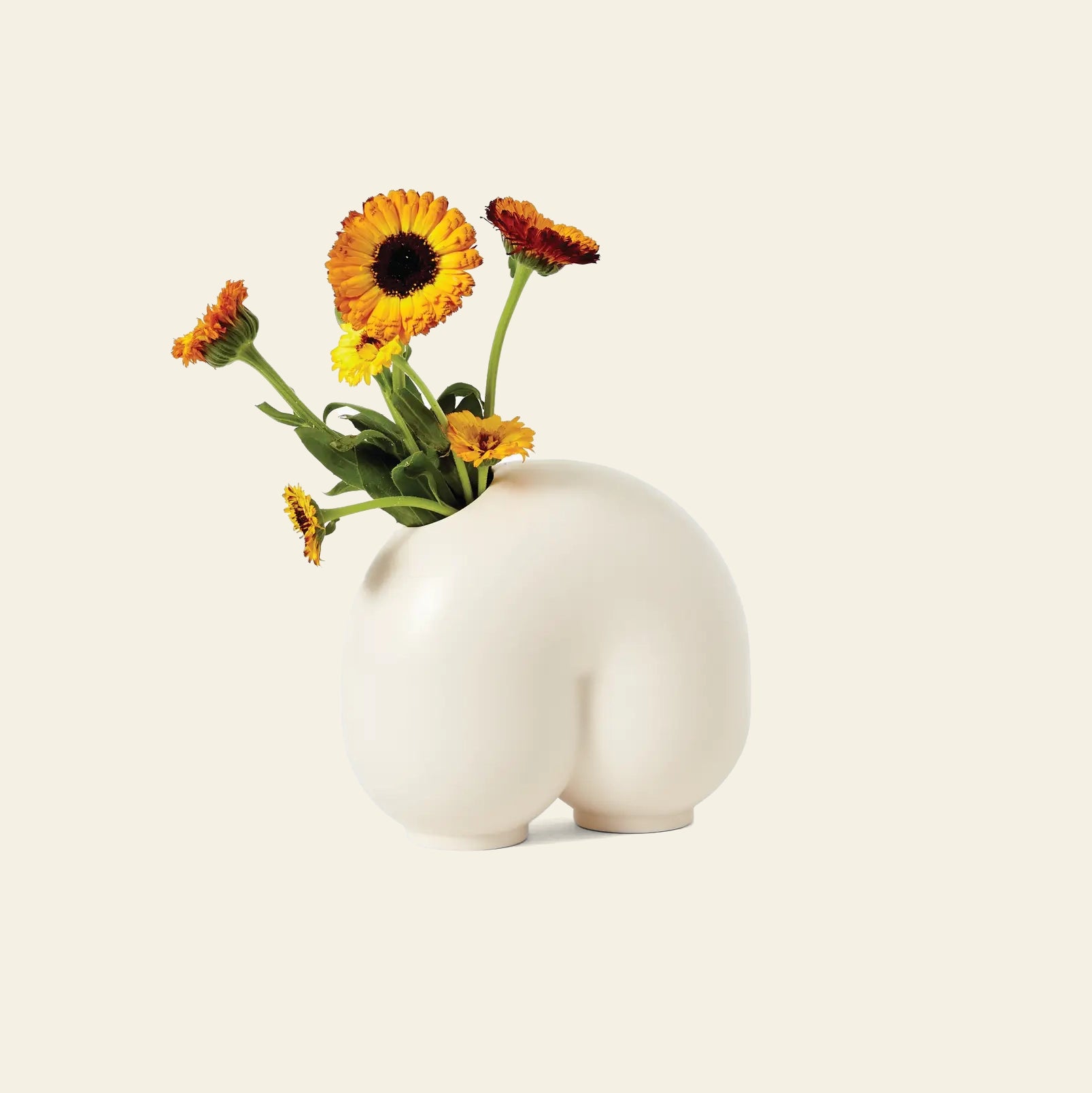 The Areaware Kirby Vase Enny by Areaware is a white ceramic vase shaped like human buttocks, perfect as a quirky best bud vase, shown holding yellow and orange flowers against a plain off-white background.
