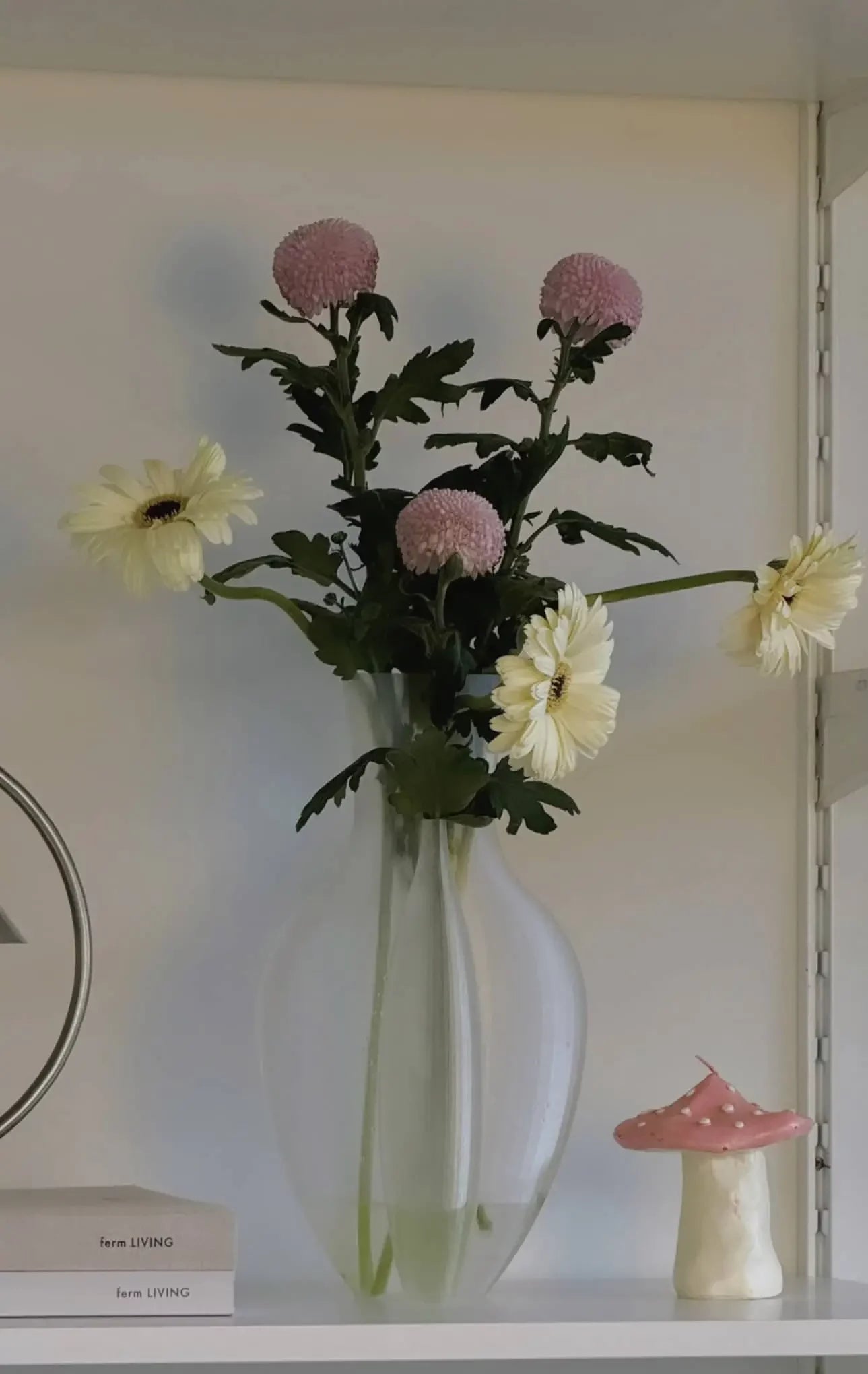 A clear Argot Studio Double Vessel by Argot Studio, inspired by the Double Vessel design, displays yellow and light purple flowers on a white shelf beside a small pink ceramic mushroom and stacked books.
