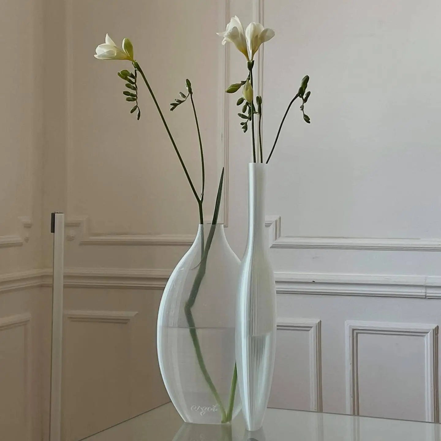 Two Argot Studio Double Vessel vases by Argot Studio, each holding a single white flower stem with buds, sit on a glass table against an elegant pale wall—a refined modern twist on classic decor.