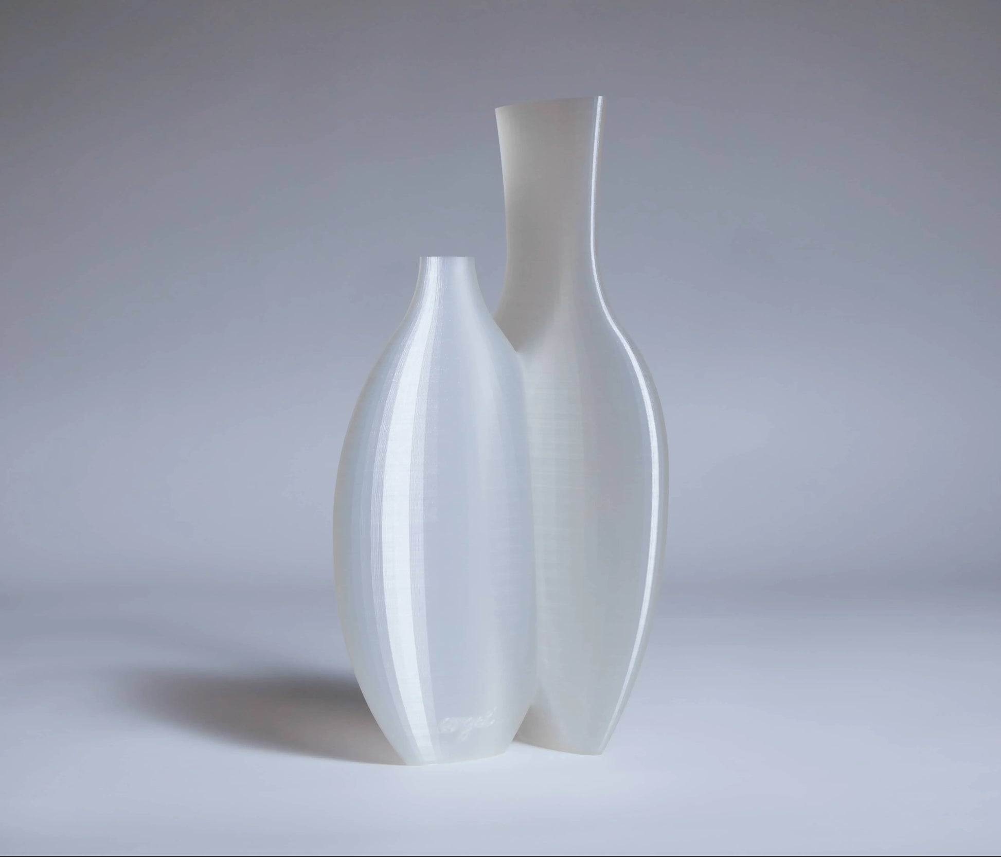 Two tall, modern, semi-translucent white vases of varying heights stand side by side on a light grey surface—a striking Argot Studio Double Vessel that reimagines classic form.