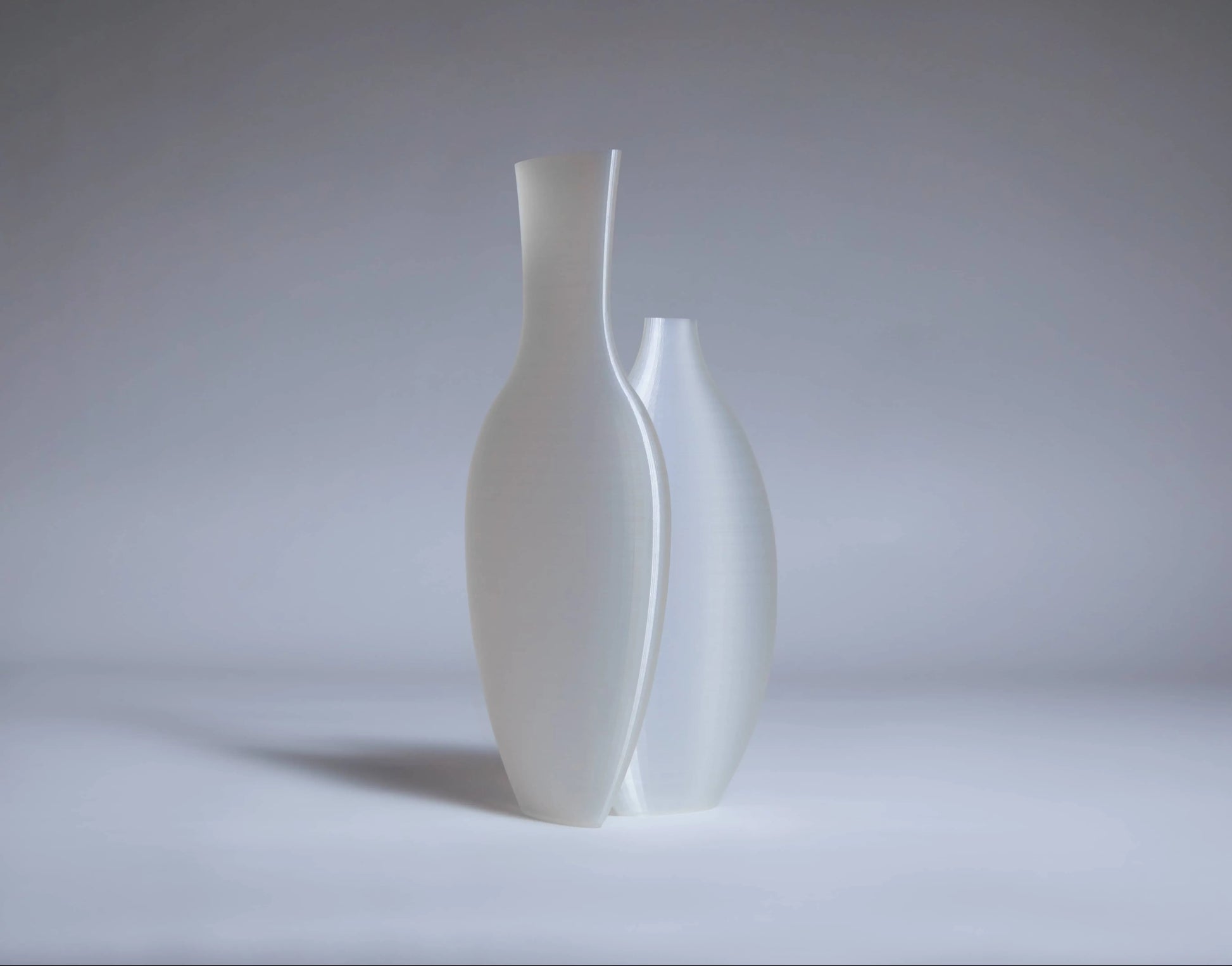 The Argot Studio Double Vessel by Argot Studio features two elegant matte white ceramic vases with elongated, curved forms—a modern take on classic shapes. One vase stands slightly taller and slimmer than the other, set against a soft gray background.