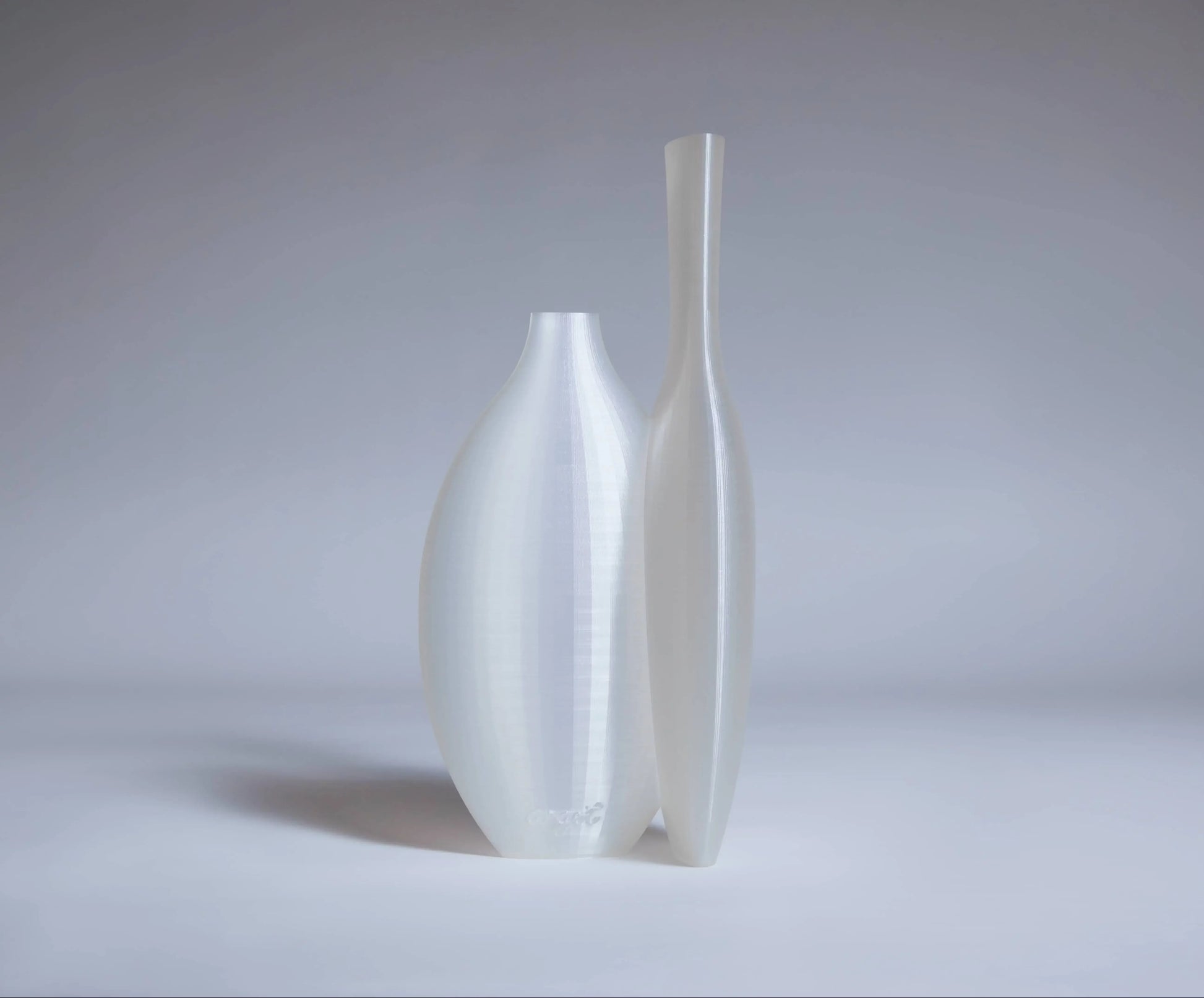 The Argot Studio Double Vessel by Argot Studio features two tall, modern white vases with smooth, minimalist forms, side by side on a light surface against a neutral gray background.