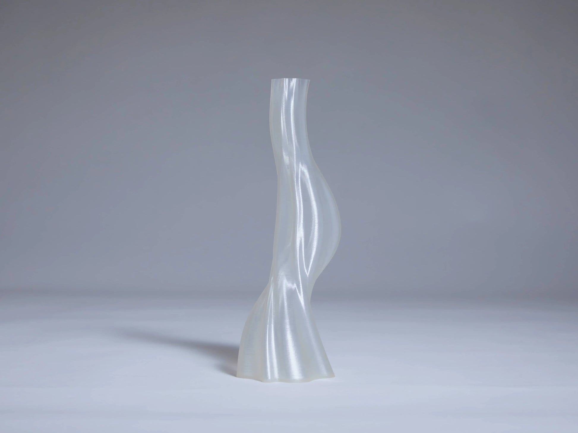 The Argot Studio Les Hortenses Vase, Small—tall, translucent, and abstract with a twisted silhouette—stands on a smooth light gray surface, offering a contemporary touch against a plain gray background.