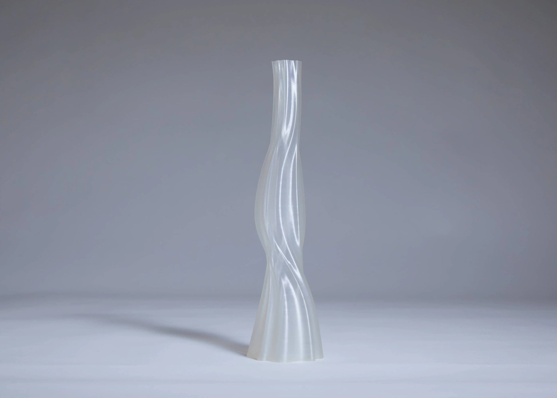 The Argot Studio Les Hortenses Vase, Tall, by Argot Studio features a slender, translucent contemporary form with a twisted sculptural design, standing on a light gray surface against a plain gray background.