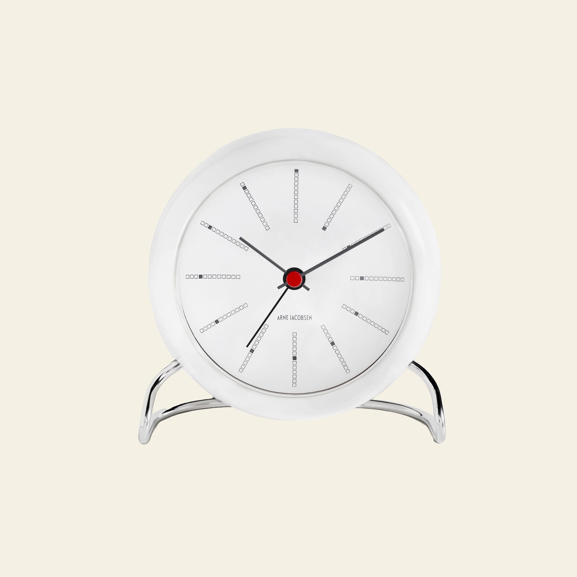 The Arne Jacobsen Bankers Table Clock 11cm by Arne Jacobsen is a modern white analog clock with a red center, black hands, and minimalist design on a silver stand, displayed against a light beige background.