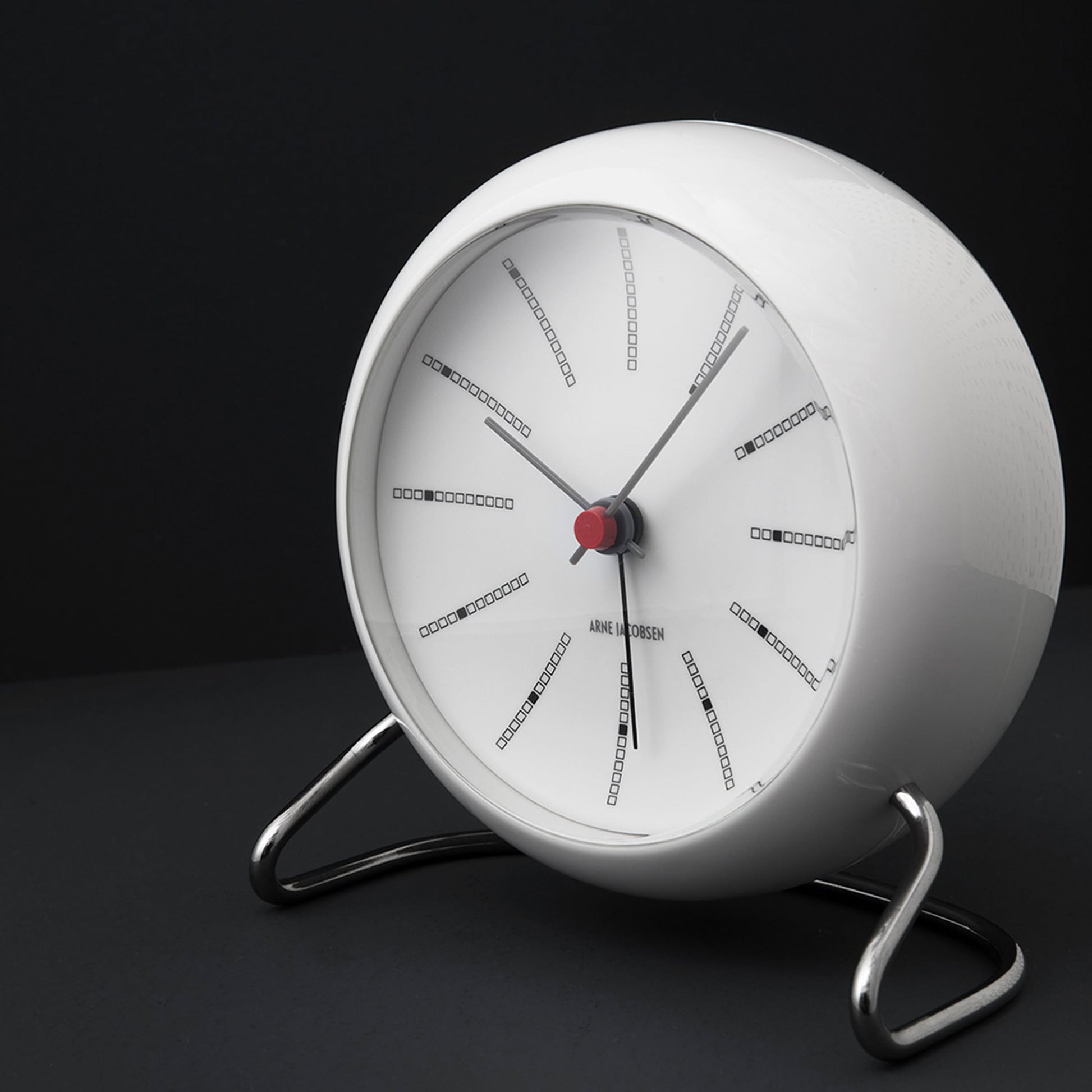 The Arne Jacobsen Bankers Table Clock 11cm by Arne Jacobsen is a modern round white clock with black hour marks, three hands, and a red center, set on a metal frame and inspired by minimalist design.