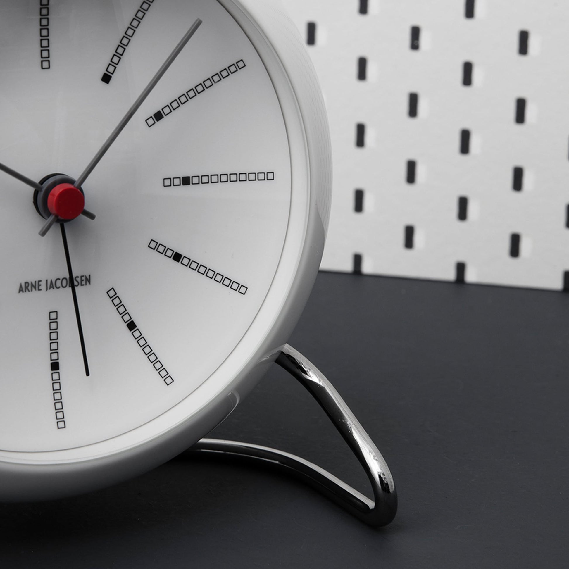 A close-up of the Arne Jacobsen Bankers Table Clock 11cm shows its modern white analog face, black and red hands, minimalist square markers, and chrome stand by Arne Jacobsen, set on a black surface with a white perforated background.
