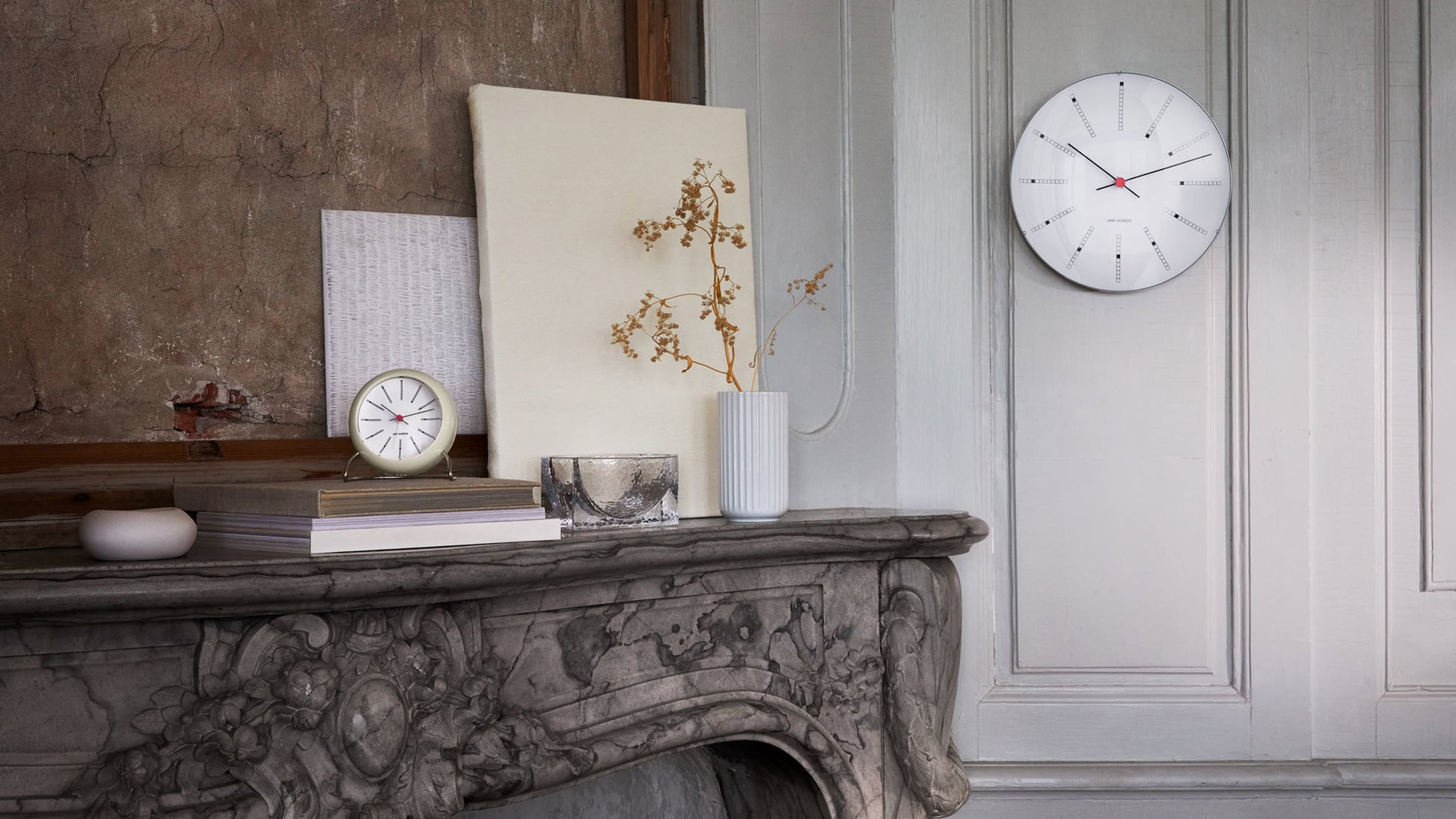 An ornate marble mantel with books, art, a vase of dried branches, a silver candle holder, and the Arne Jacobsen Bankers Table Clock 11cm by Arne Jacobsen sits against a textured wall; nearby is a minimalist white clock on a light gray paneled wall.