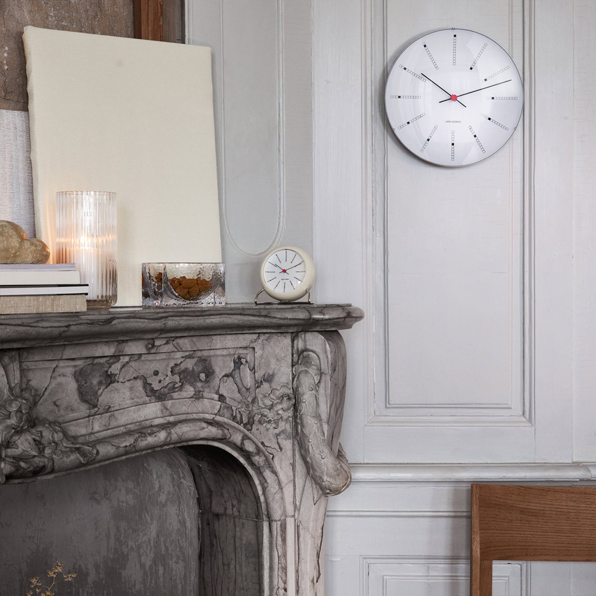 A marble fireplace with decorative carvings holds books, a glass of water, a candle, and the Arne Jacobsen Bankers Table Clock 11cm. Above, a modern wall clock hangs on the paneled wall of this softly lit, minimalist-inspired room.