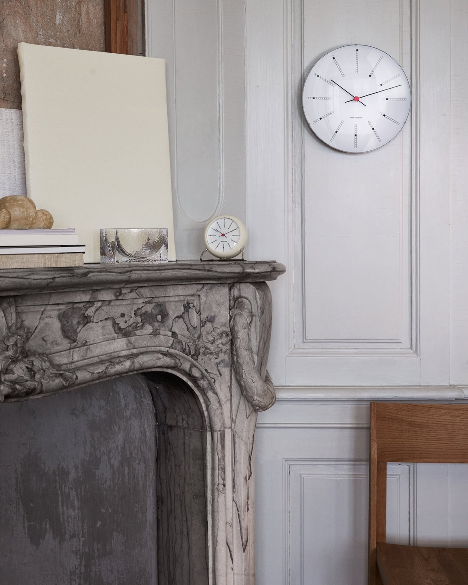 A marble fireplace displays books, an Arne Jacobsen Bankers Table Clock 11cm, and a blank canvas on the mantel. Above, a round minimalist wall clock hangs on paneled walls, with part of a wooden chair to the right.