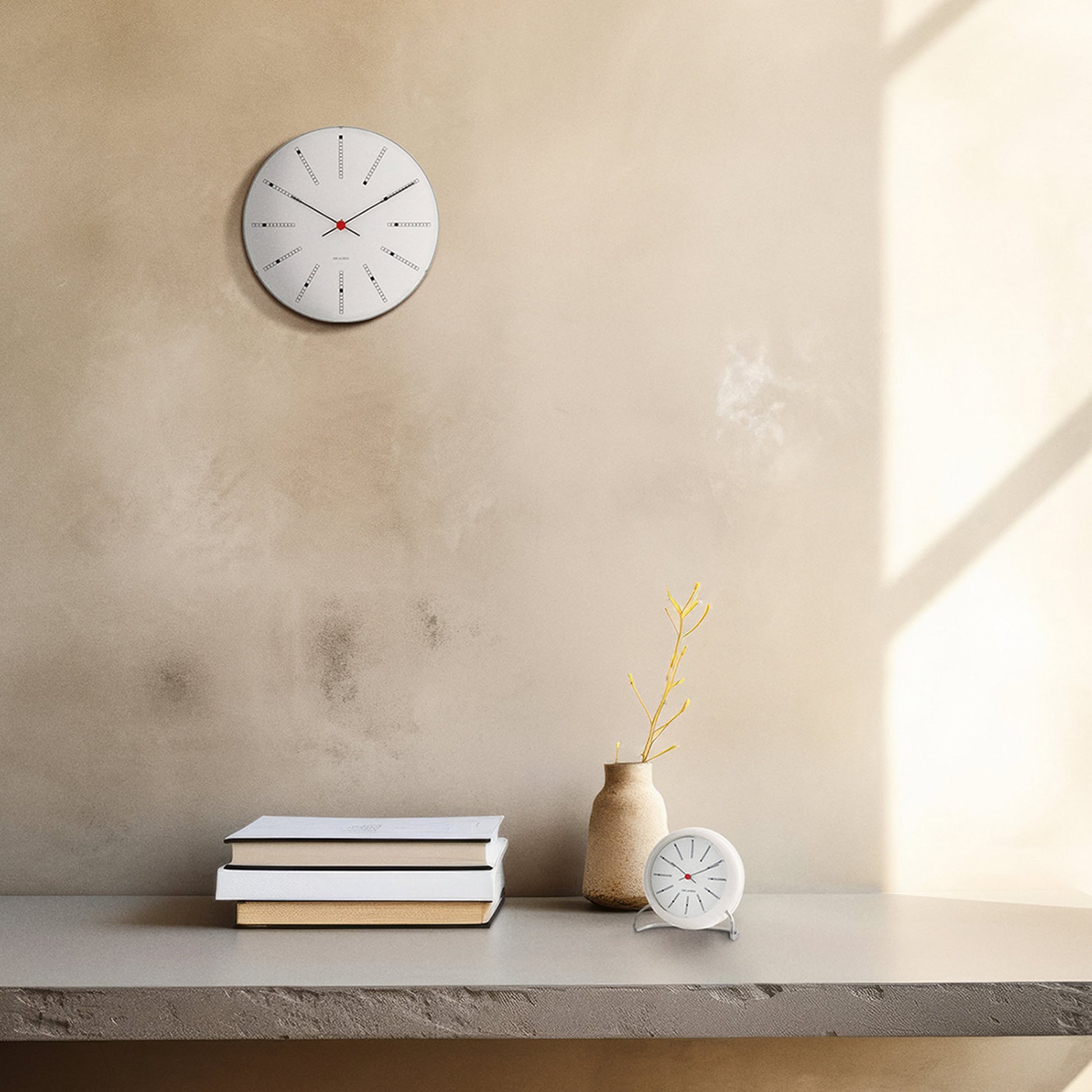 A minimalist room features a round wall clock, books, the Arne Jacobsen Bankers Table Clock 11cm by Arne Jacobsen, and a beige vase with a yellow branch on a light gray surface against a textured beige wall dappled with sunlight.