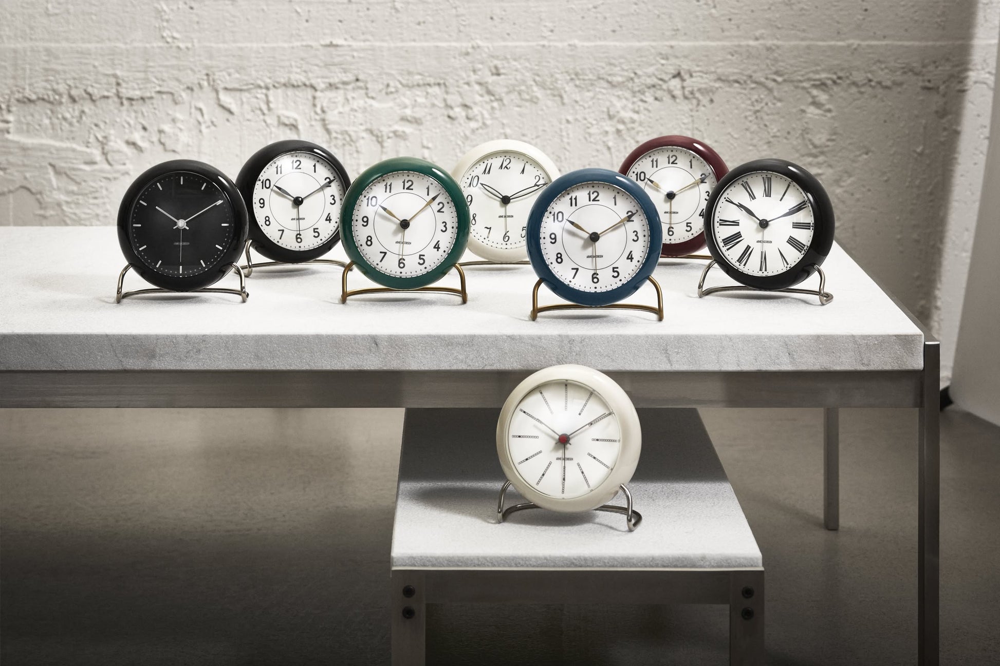 Eight Arne Jacobsen Bankers Table Clock 11cm by Arne Jacobsen, each with a unique colored frame and minimalist design, are arranged on two white tables—seven above, one below—set against a textured white wall.