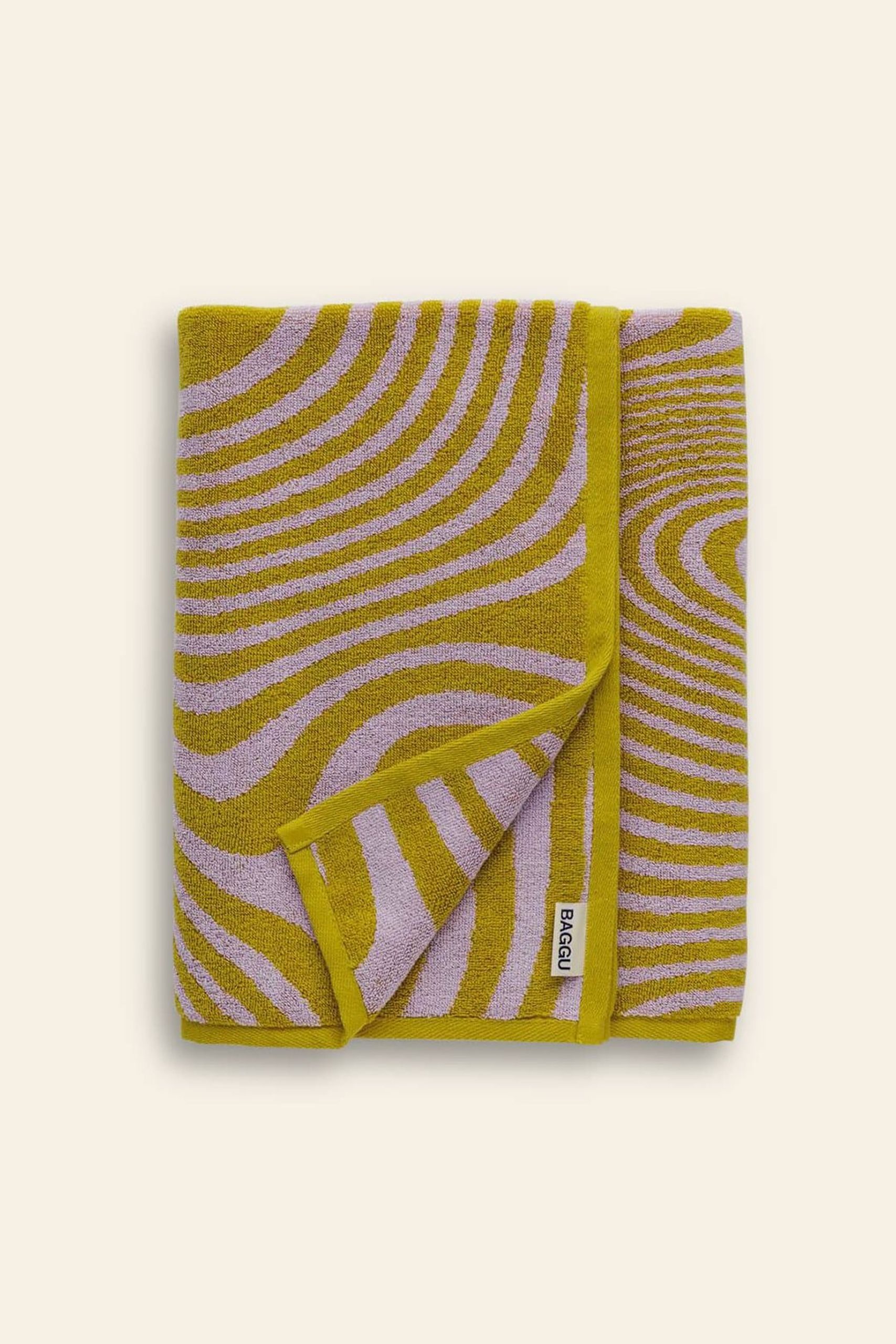 A BAGGU Bath Towel by BAGGU, folded to reveal its yellow and lavender wavy line pattern and a white edge tag with the brand name, is displayed on a light beige background.