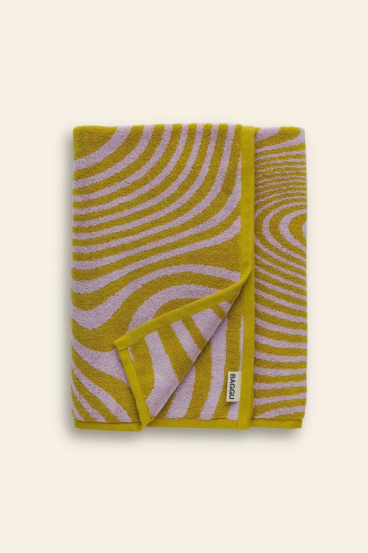 A folded BAGGU Bath Towel by BAGGU, featuring a vibrant wavy mustard yellow and lavender abstract print with a small white brand label on the edge, set against a plain light beige background—ideal as a sunflower-inspired bath towel accent.