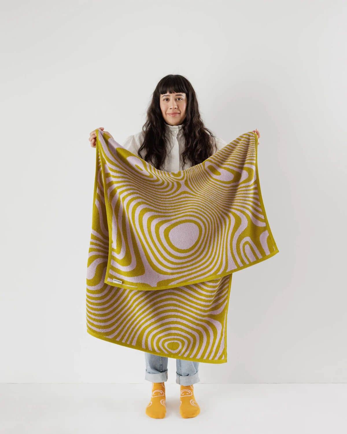 A woman with long dark hair holds up the BAGGU Bath Towel by BAGGU, featuring yellow and beige geometric patterns. She stands against a plain white background, dressed in a white shirt, light blue jeans, and yellow socks.