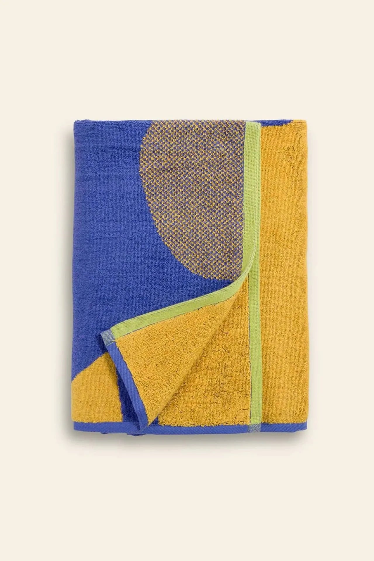 The BAGGU Bath Towel features bold blue and mustard sections, a large textured brown circle, and green trim. Displayed on a plain light background, this vibrant towel is crafted by BAGGU.