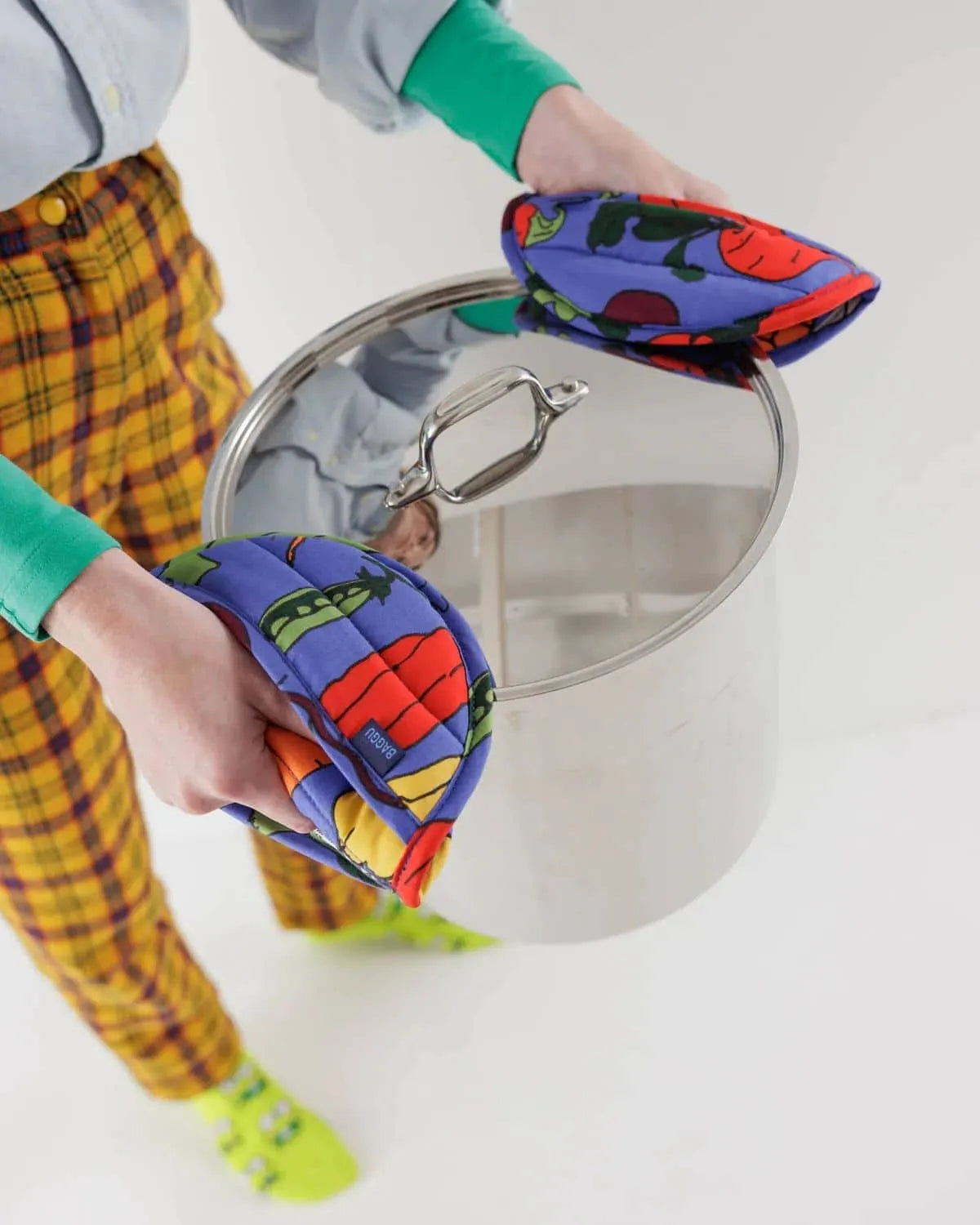 Wearing yellow plaid pants and green socks, someone holds a large stainless steel pot with a lid using BAGGU Pot Holder Set of 2 by BAGGU—psychedelic organic cotton pot holders with red, blue, and green patterns.