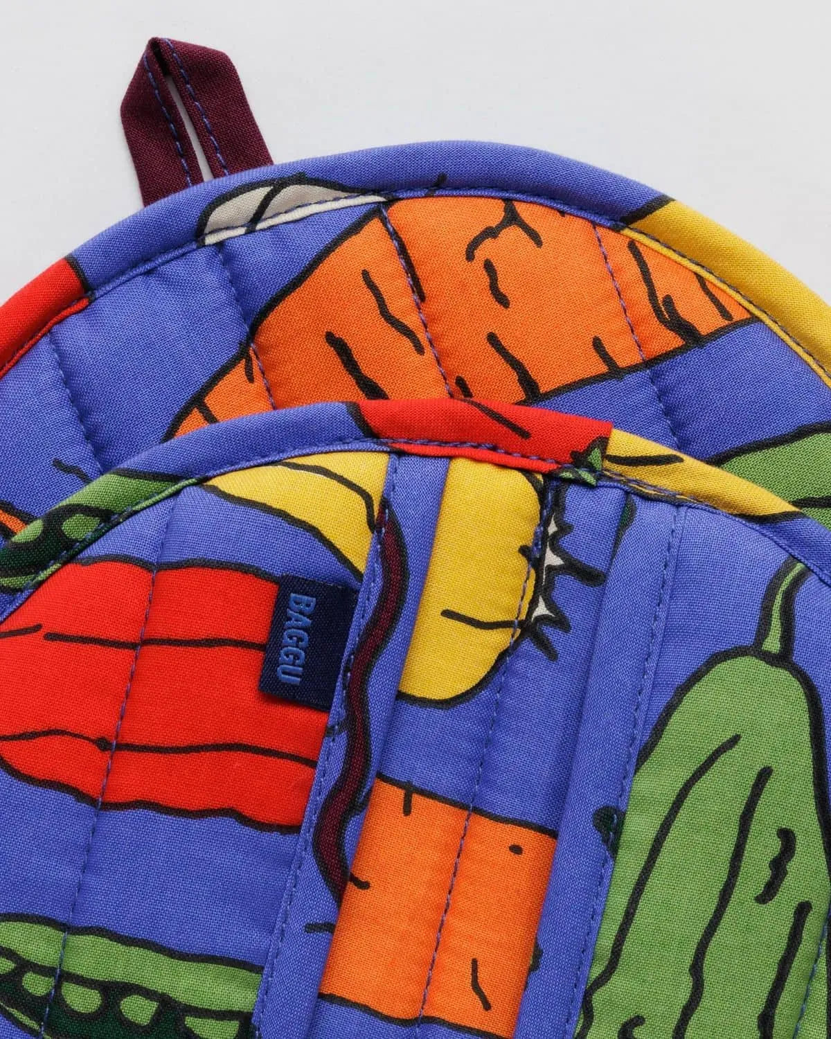 Close-up of the BAGGU Pot Holder Set of 2, made from organic cotton with colorful vegetable prints on a purple background. Features carrots, peppers, and cucumbers in a playful design and includes a navy blue BAGGU tag.