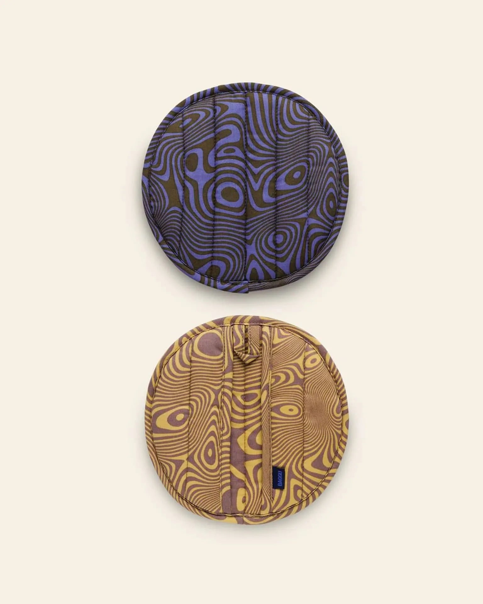 The BAGGU Pot Holder Set of 2 by BAGGU features round designs with abstract wavy patterns—one in blue/black, one in yellow/brown—crafted from organic cotton with zippers and tags, shown on a plain beige background.